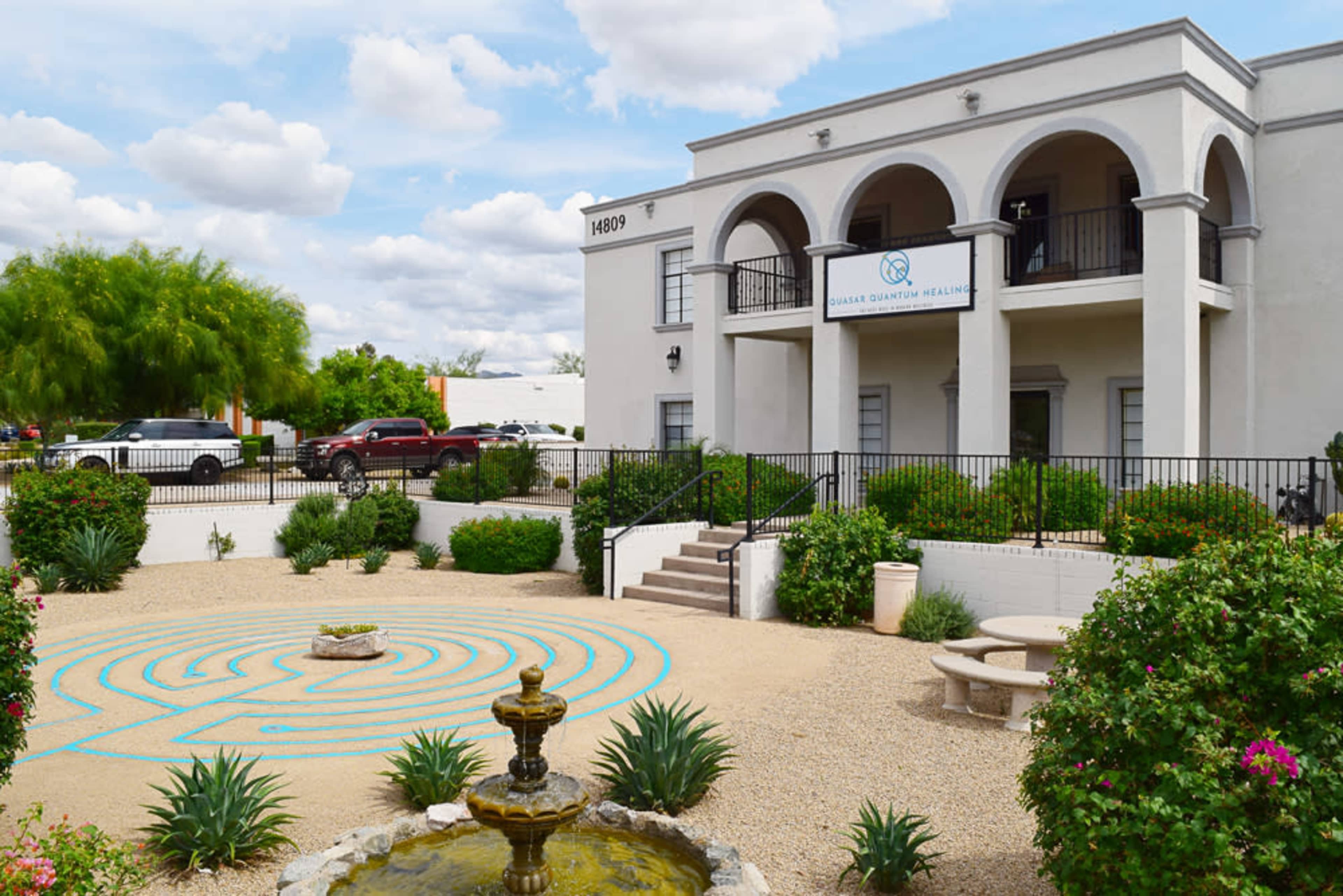 A two-story building with a sign that reads "Gisela Quantum Healing," surrounded by landscaped gardens featuring a fountain and a blue-painted labyrinth in the foreground.