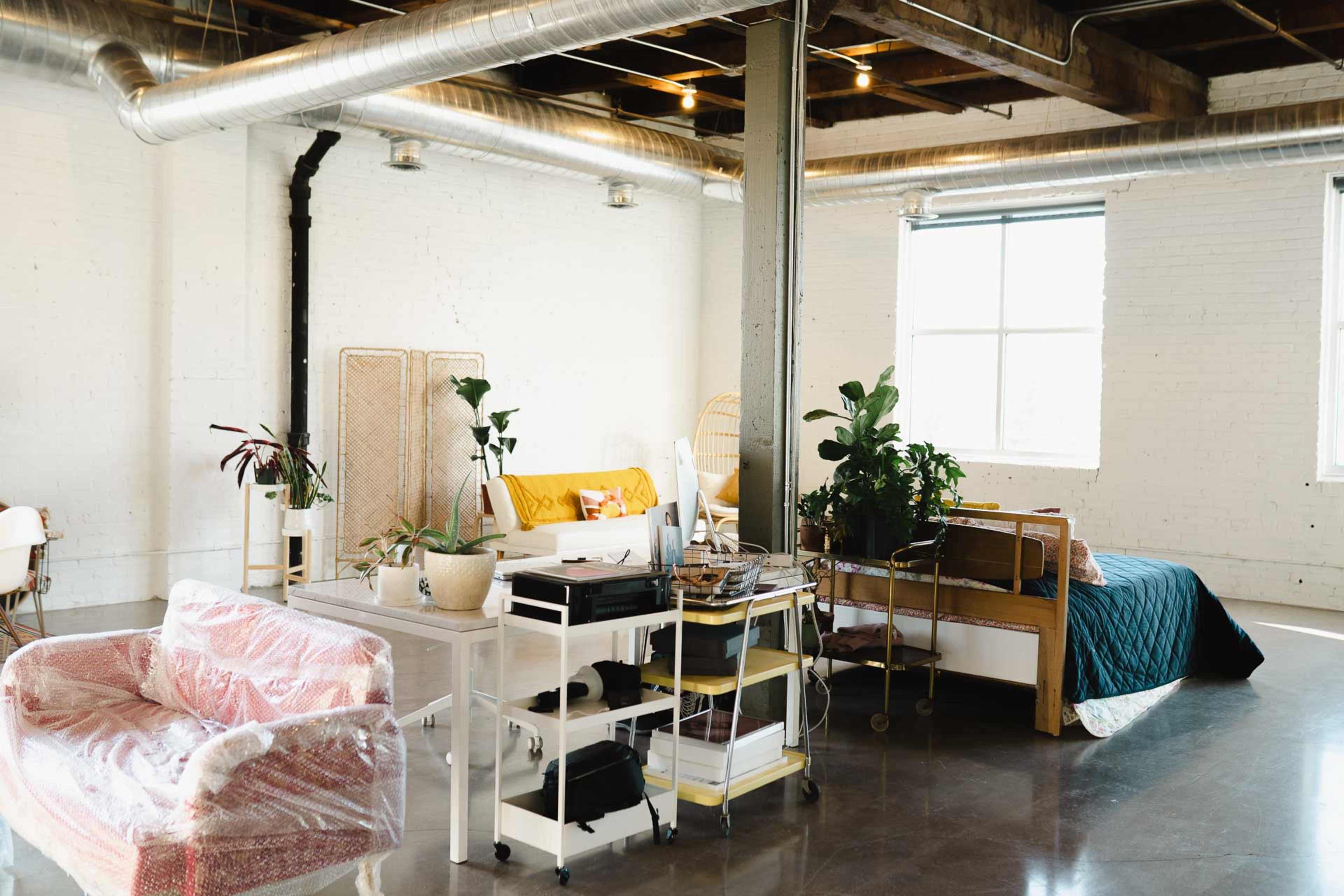 The image shows a spacious, modern loft with a seating area featuring a patterned sofa, a plant-filled workspace, and a bed adorned with colorful bedding.