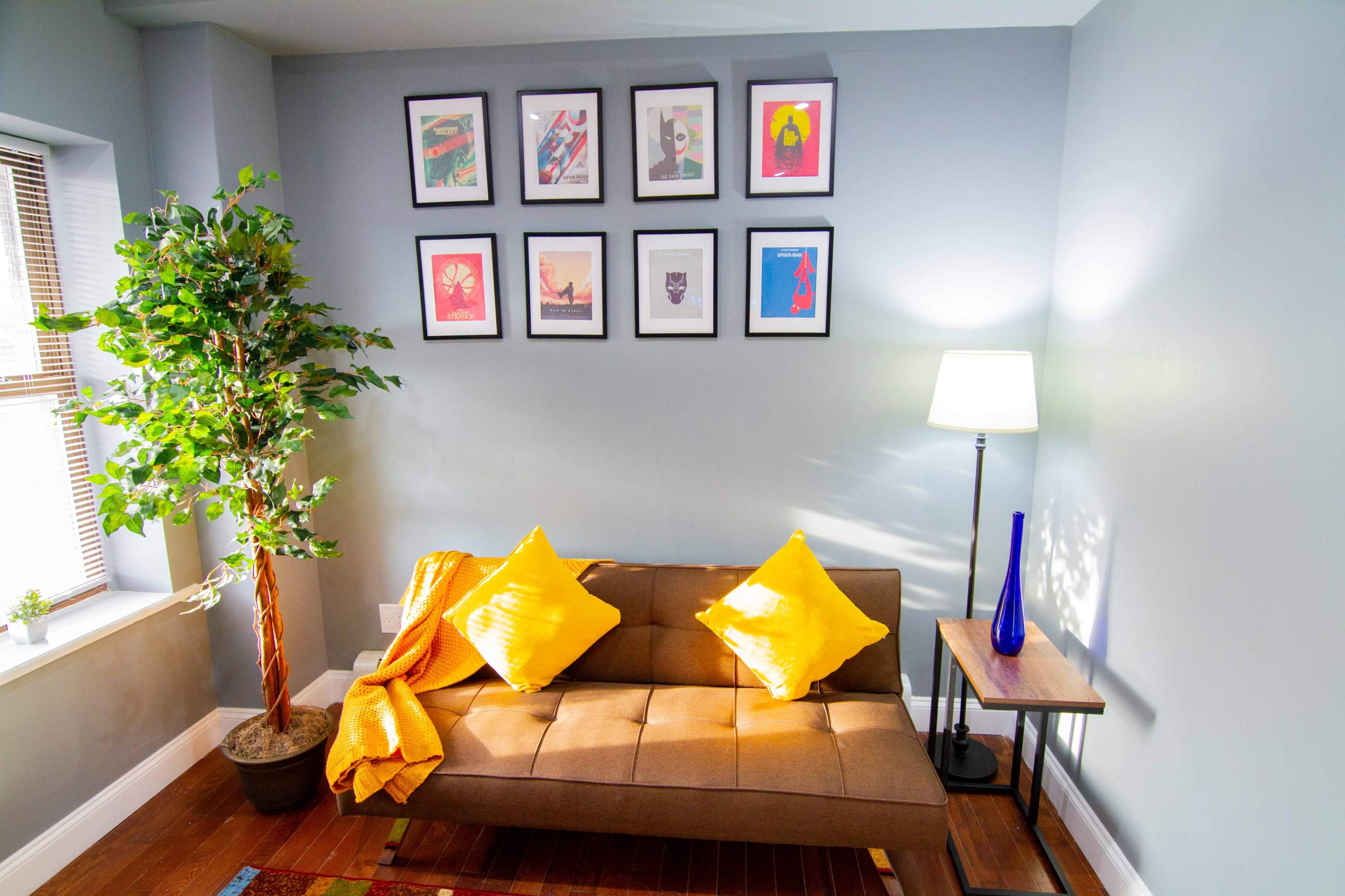 A cozy living room corner features a brown sofa with yellow cushions, a standing lamp, a side table, an indoor plant, and framed artwork on the wall.