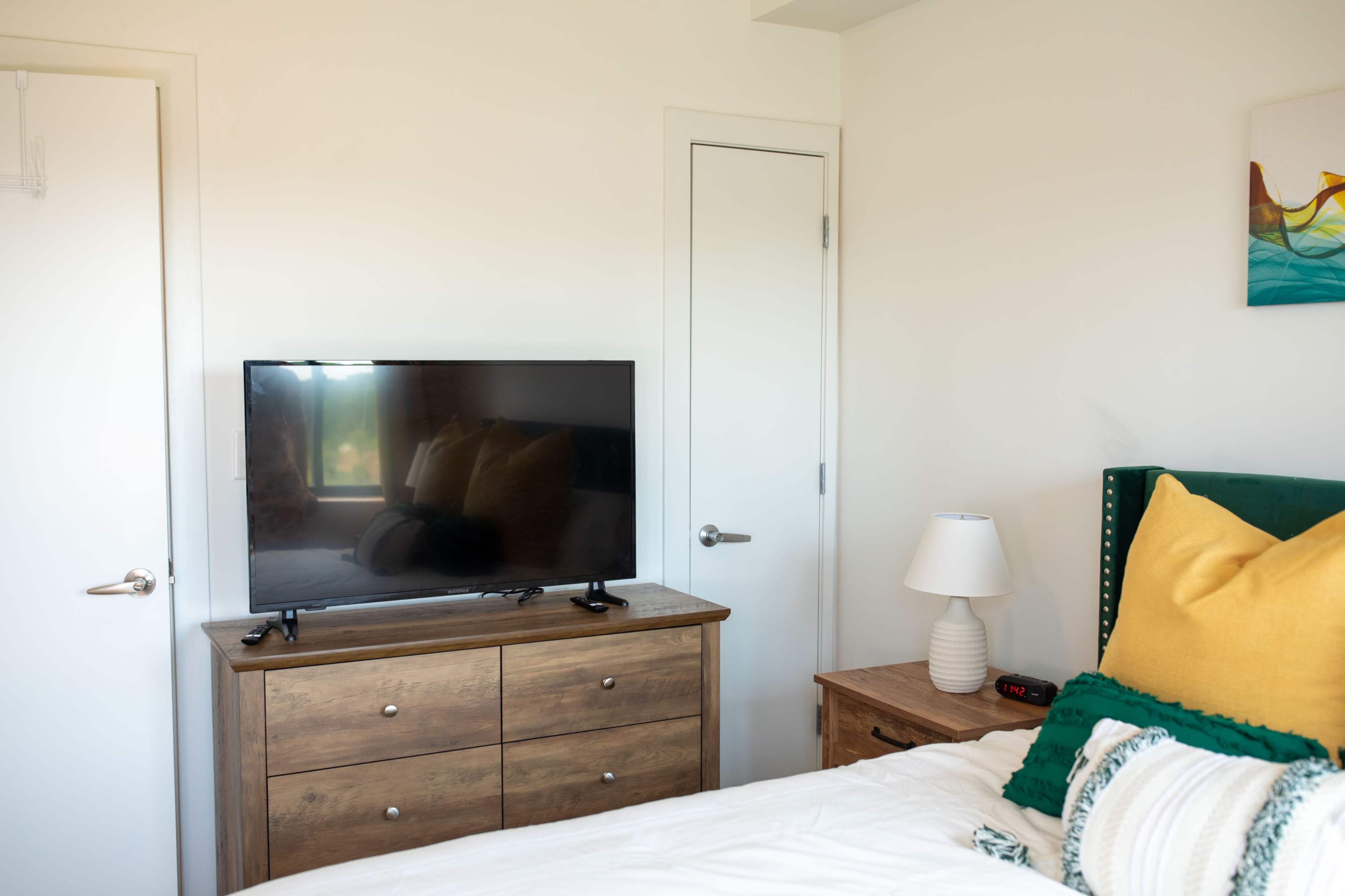The image shows a cozy bedroom with a television on a wooden dresser, a bedside lamp, and a green upholstered headboard.