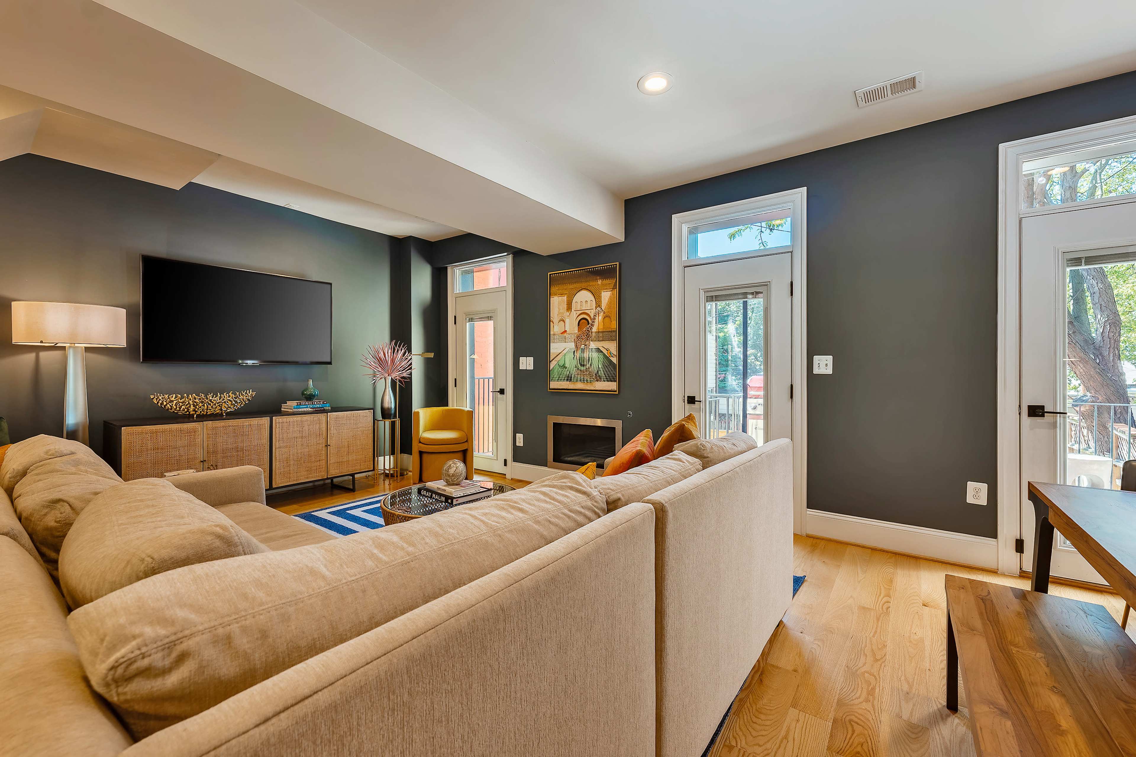 A living room features a beige sofa, a wooden media console, and large windows allowing natural light, with a modern television mounted on the wall.