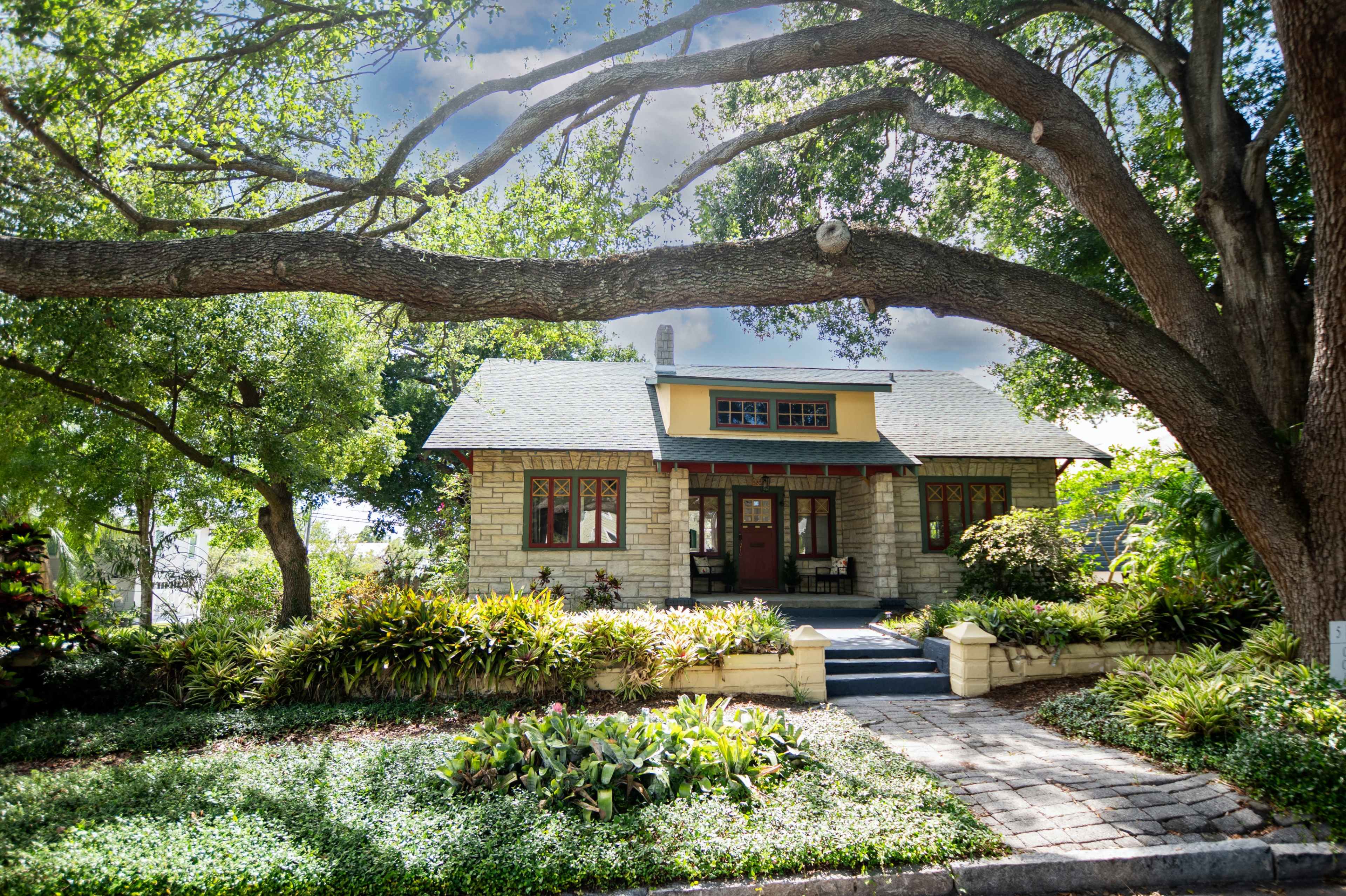 A stone cottage with a front porch is surrounded by lush greenery and large trees.