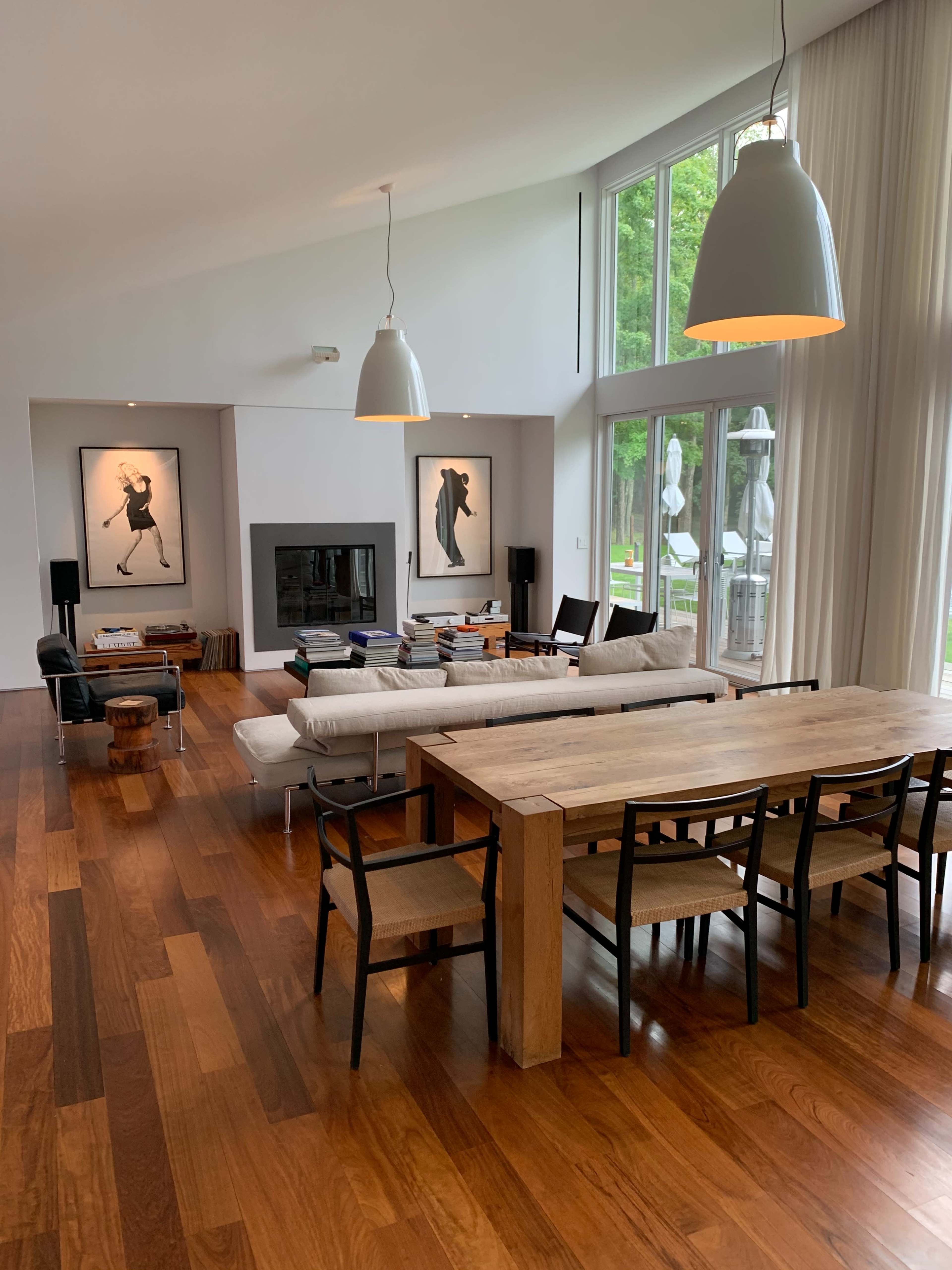 A modern living room with a wooden dining table, a white sofa, two framed artworks on the wall, and a large window letting in natural light.