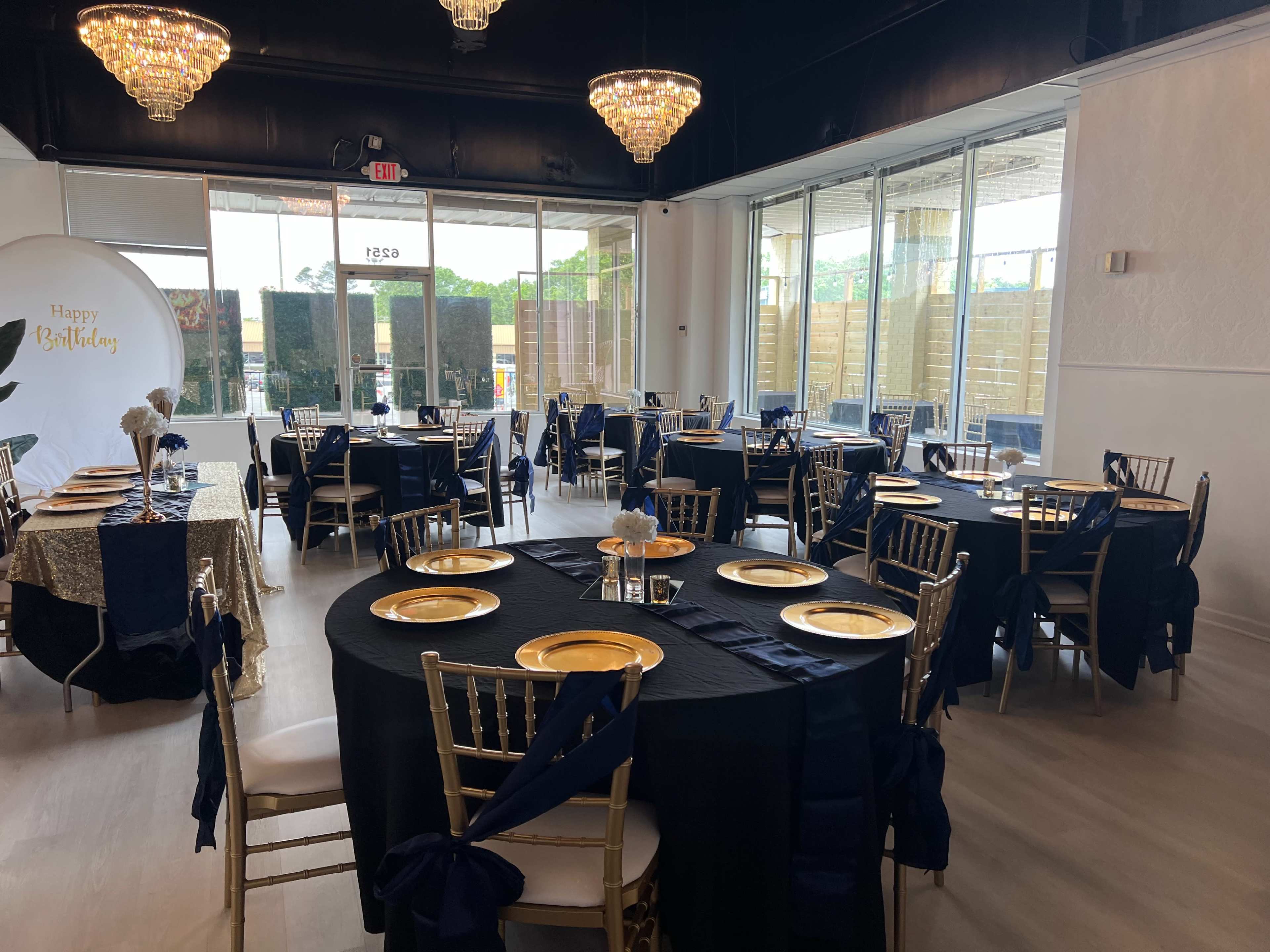 A spacious dining area is set up for a birthday celebration, featuring black tablecloths with gold plates and chiavari chairs at elegantly arranged tables.