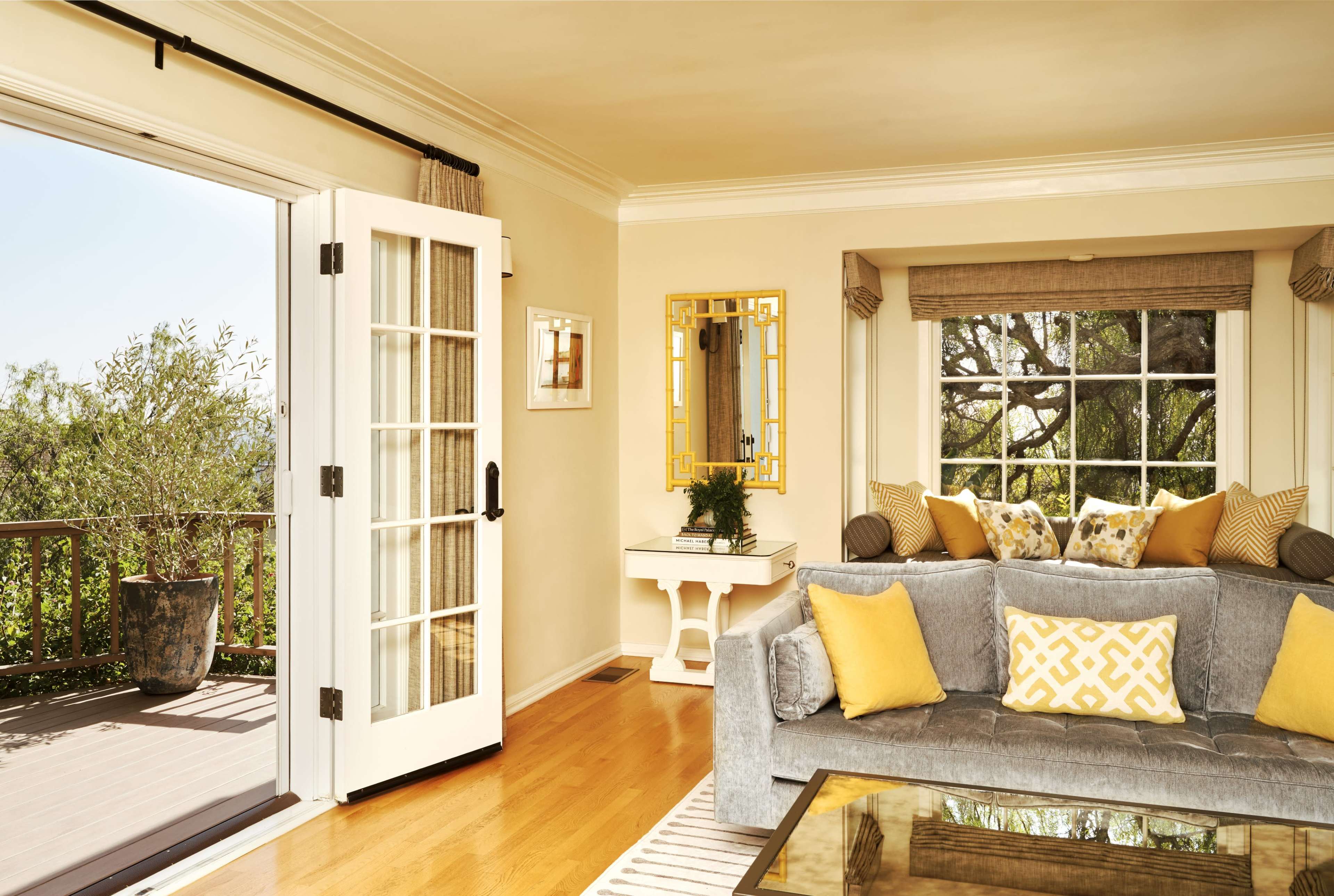 A living room features a gray couch adorned with yellow pillows, a glass-top coffee table, and French doors that open to a balcony with scenic views.