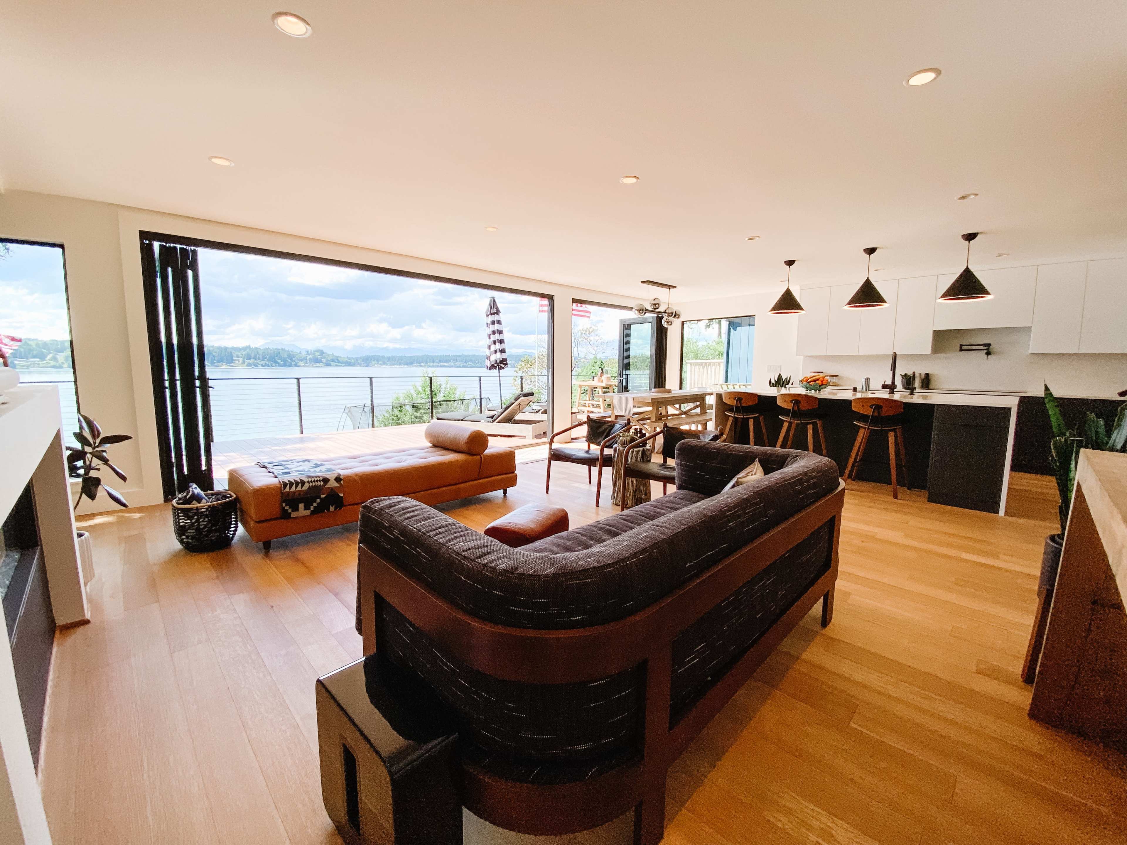 A modern living space with a large window overlooking a lake, featuring a leather couch, dining area, and a deck with outdoor seating.