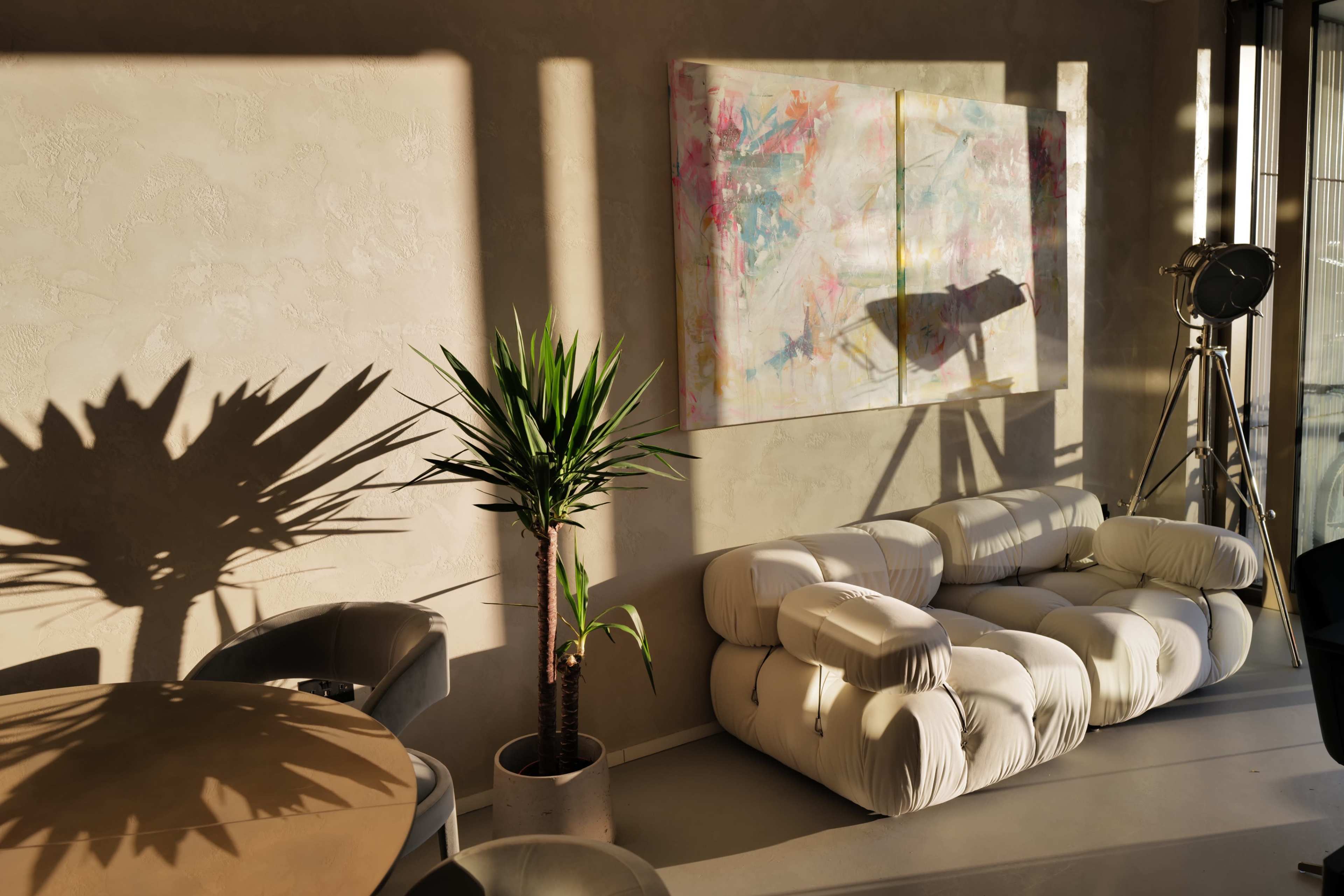 A modern living space featuring a white sofa, a round table, a potted plant, and a colorful abstract painting on the wall, with sunlight casting shadows across the room.