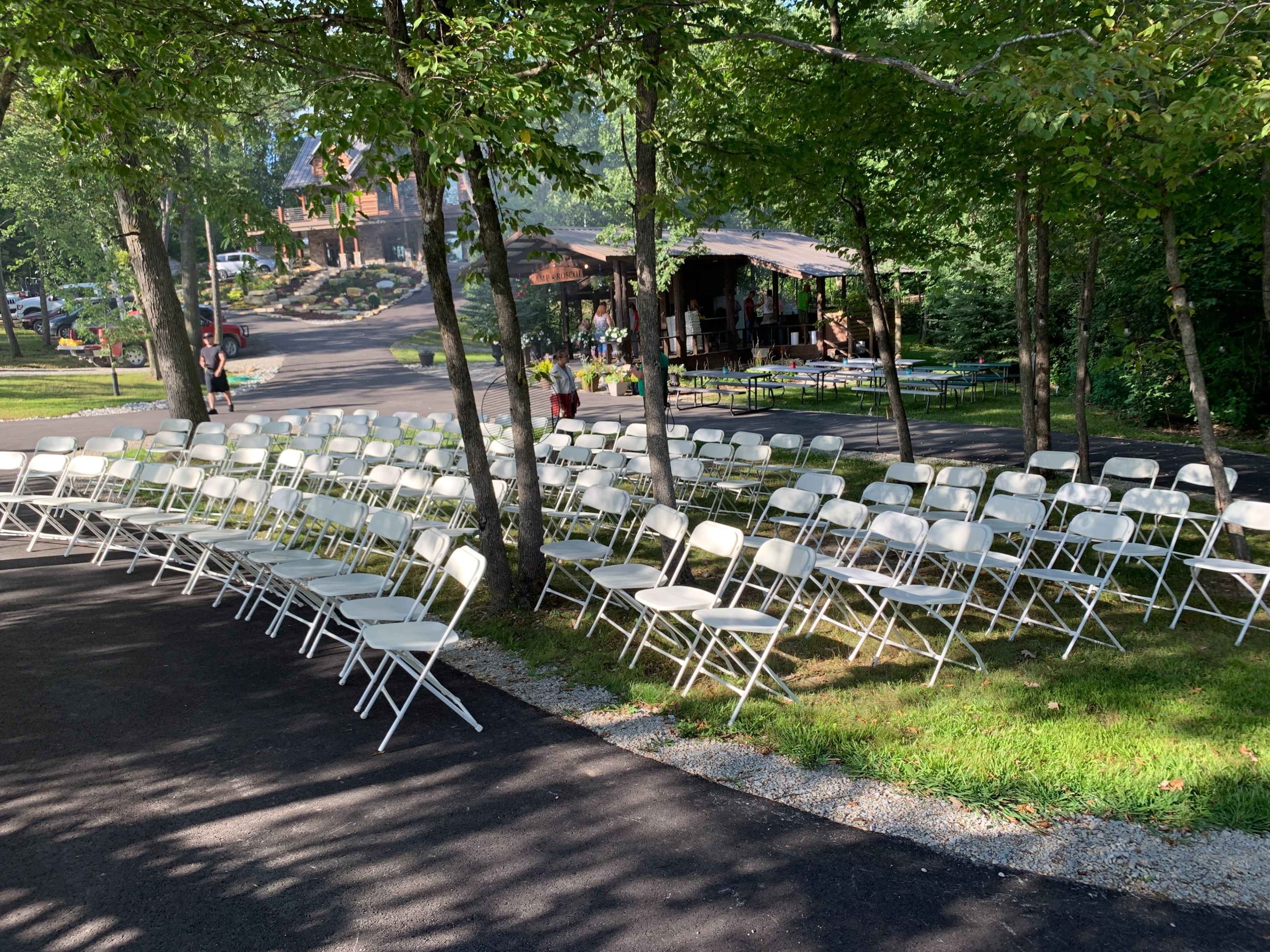 A row of white folding chairs is neatly arranged under trees in a grassy area near a winding path.