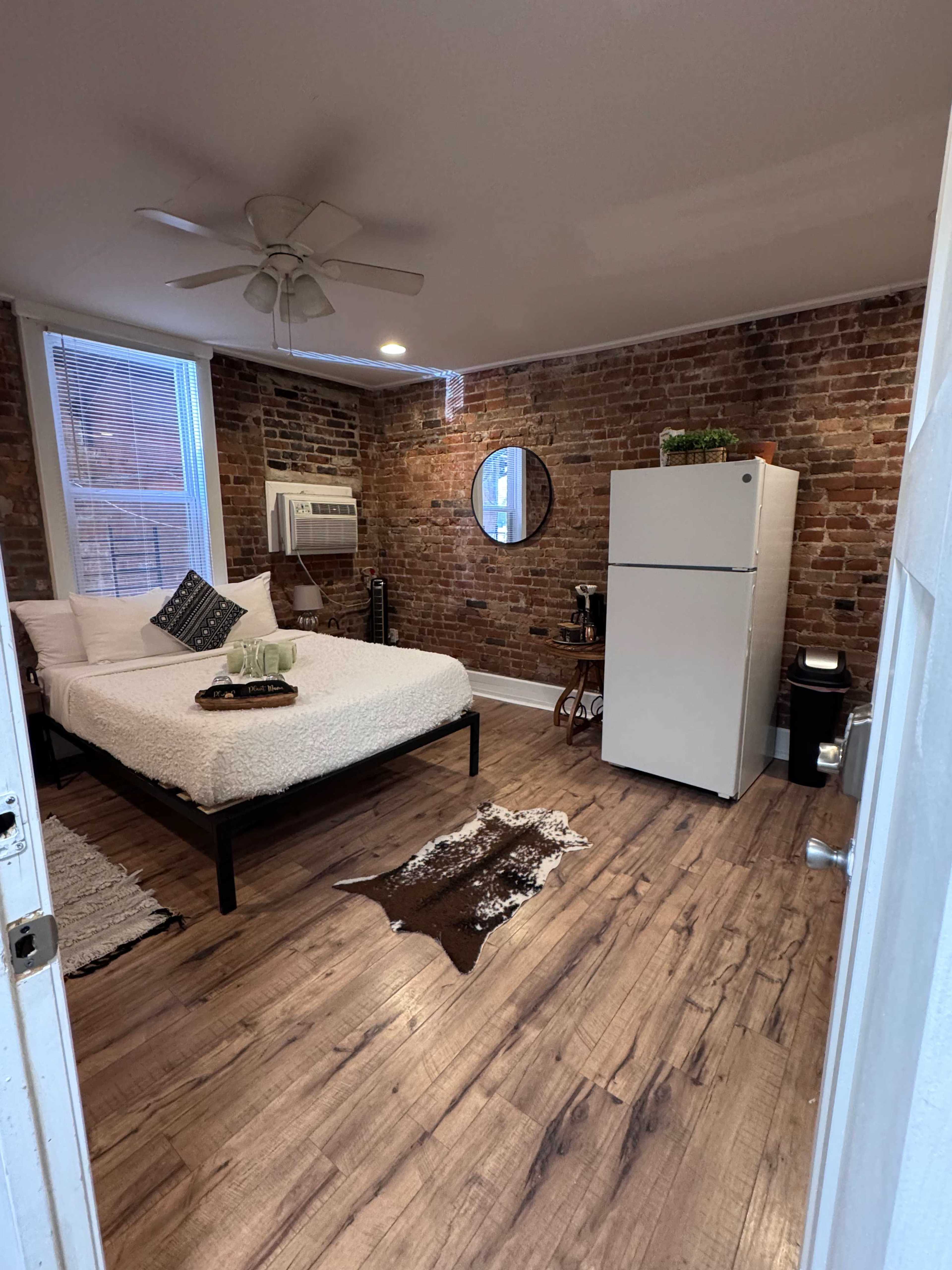 The room features a bed with white linens, a white refrigerator, and exposed brick walls, along with a small seating area.