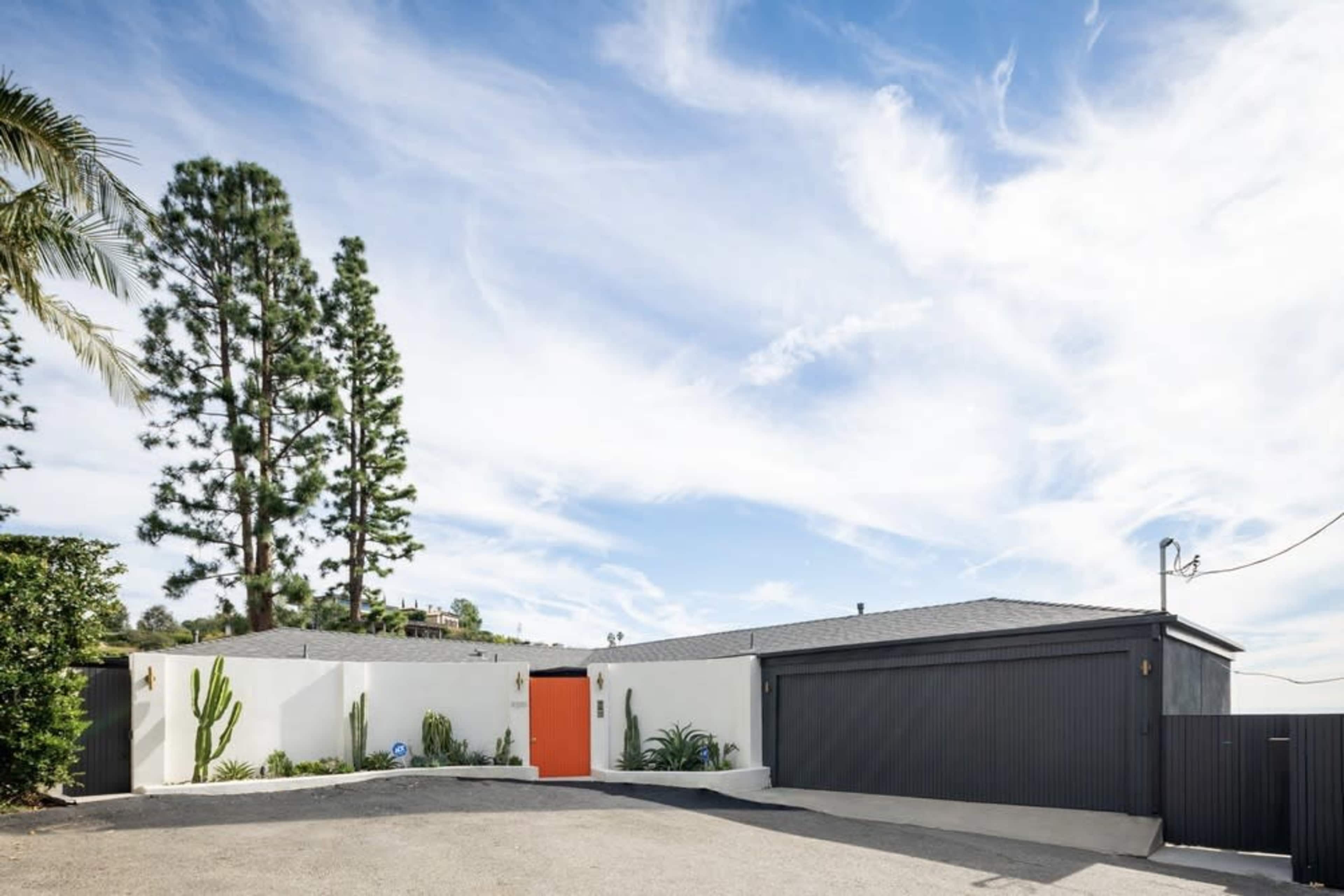 A modern house with a striking orange front door is set against a backdrop of tall pine trees and a blue sky.