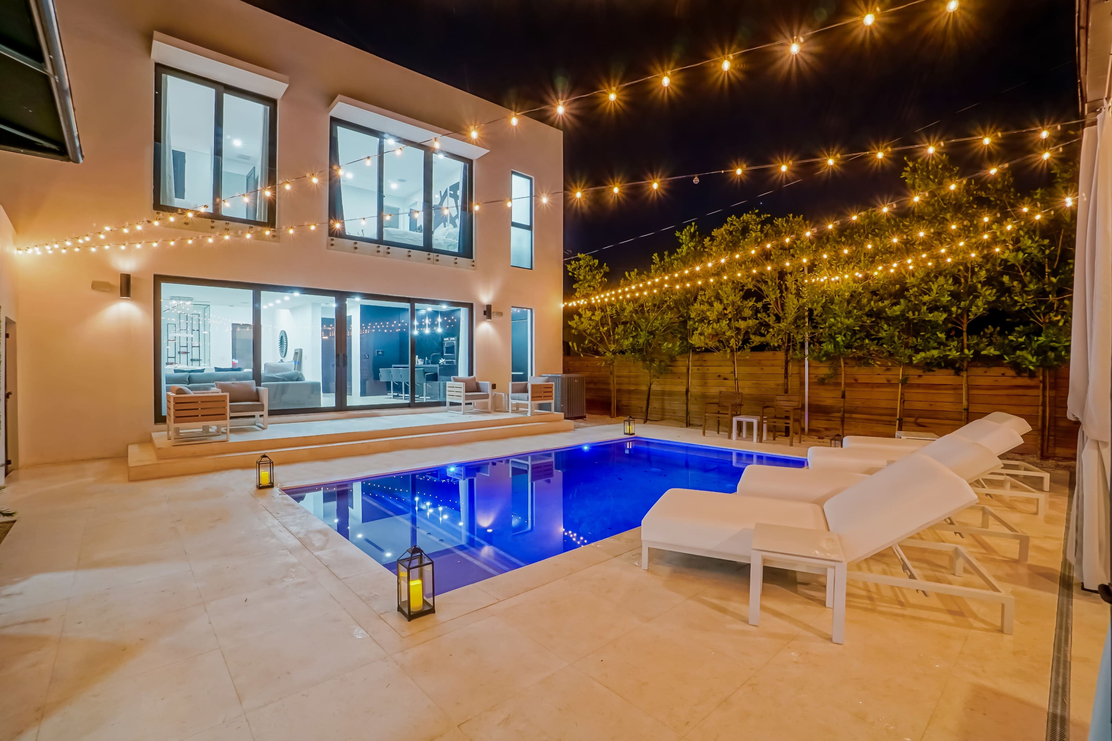 The image shows a modern backyard featuring a swimming pool illuminated by string lights, with a spacious patio and several lounge chairs.