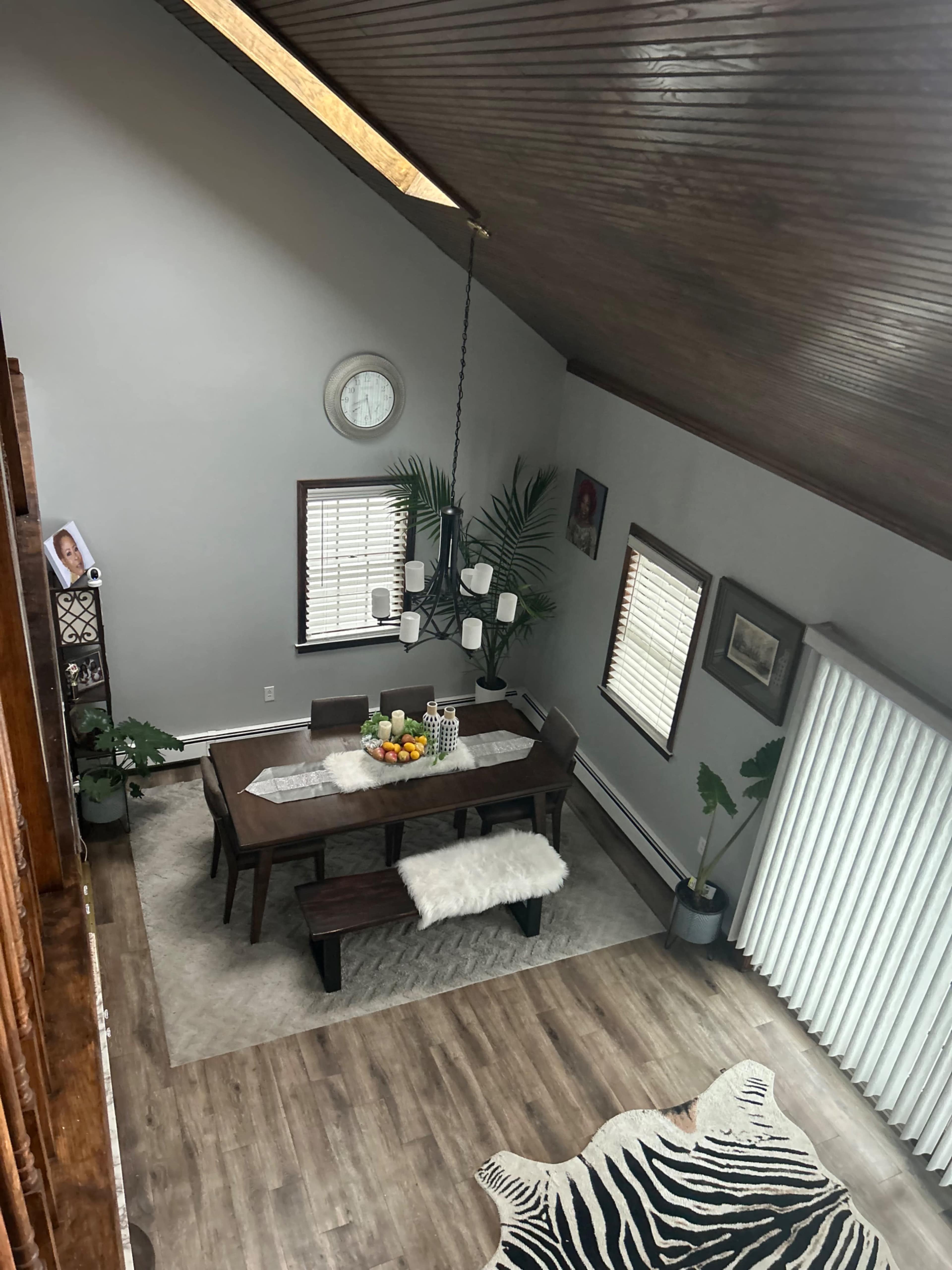 The image shows a spacious dining area featuring a wooden table surrounded by benches, with fruit on the table, large windows, and a clock on the wall.