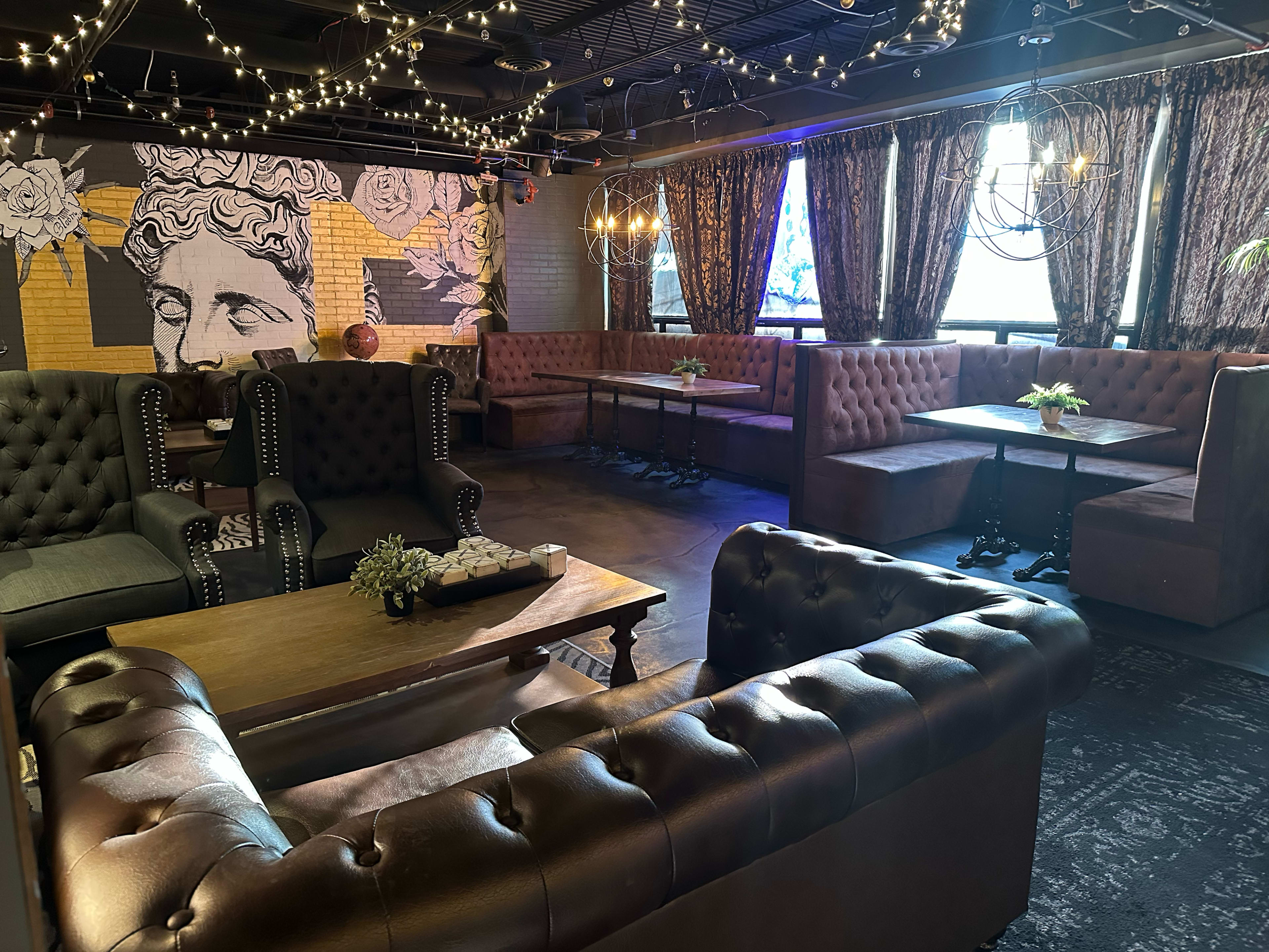 The image shows an upscale lounge featuring dark leather seating, wooden tables, and decorative lighting against a backdrop of artistic wall murals.