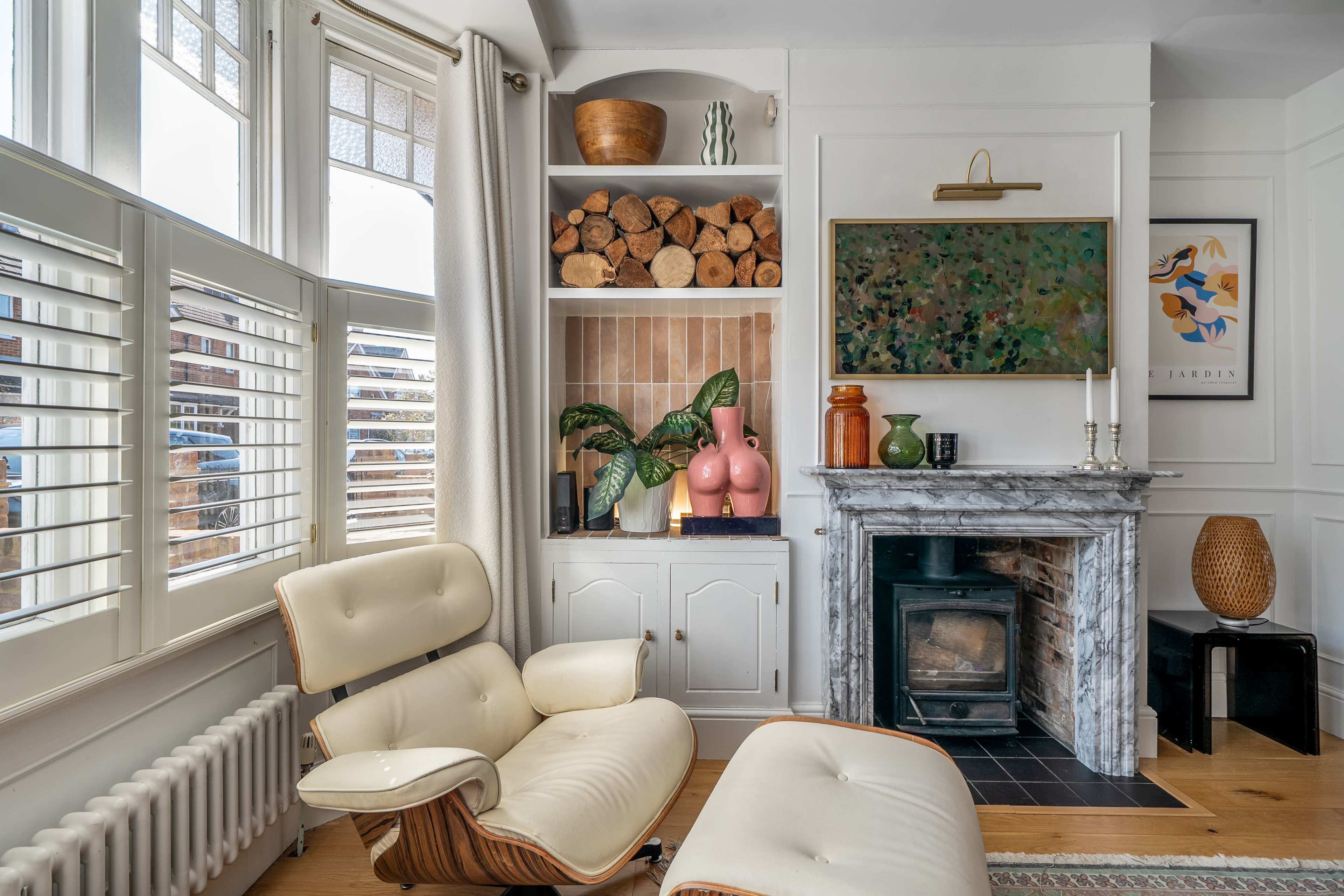 The image shows a cozy living room with a modern armchair, a marble fireplace, and shelves containing neatly stacked logs and decorative items.
