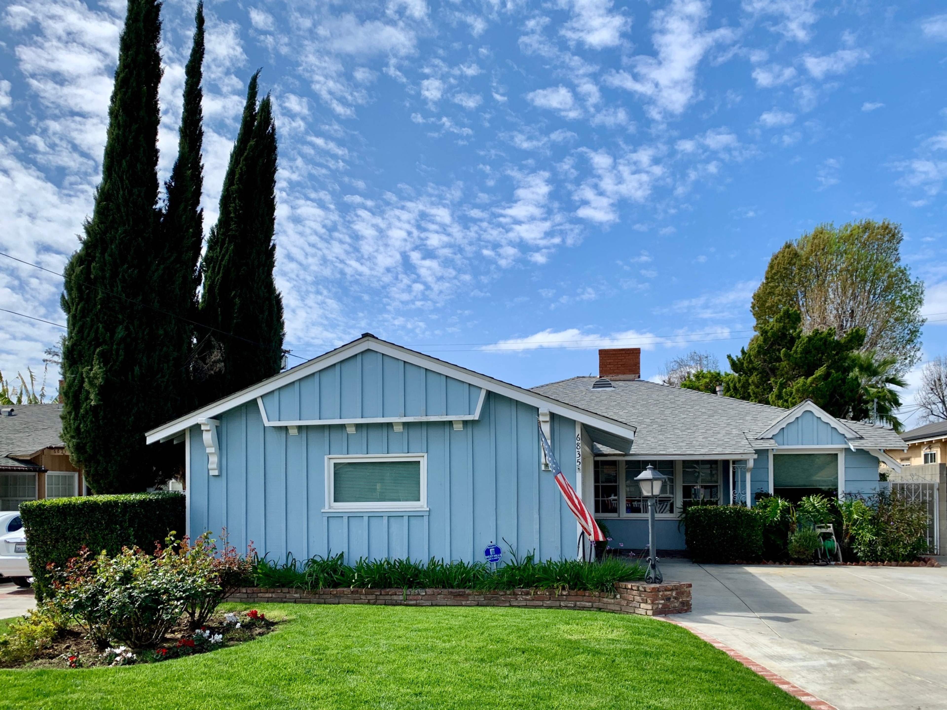 A light blue house with a front porch and an American flag is surrounded by manicured lawns and tall cypress trees under a partly cloudy sky.
