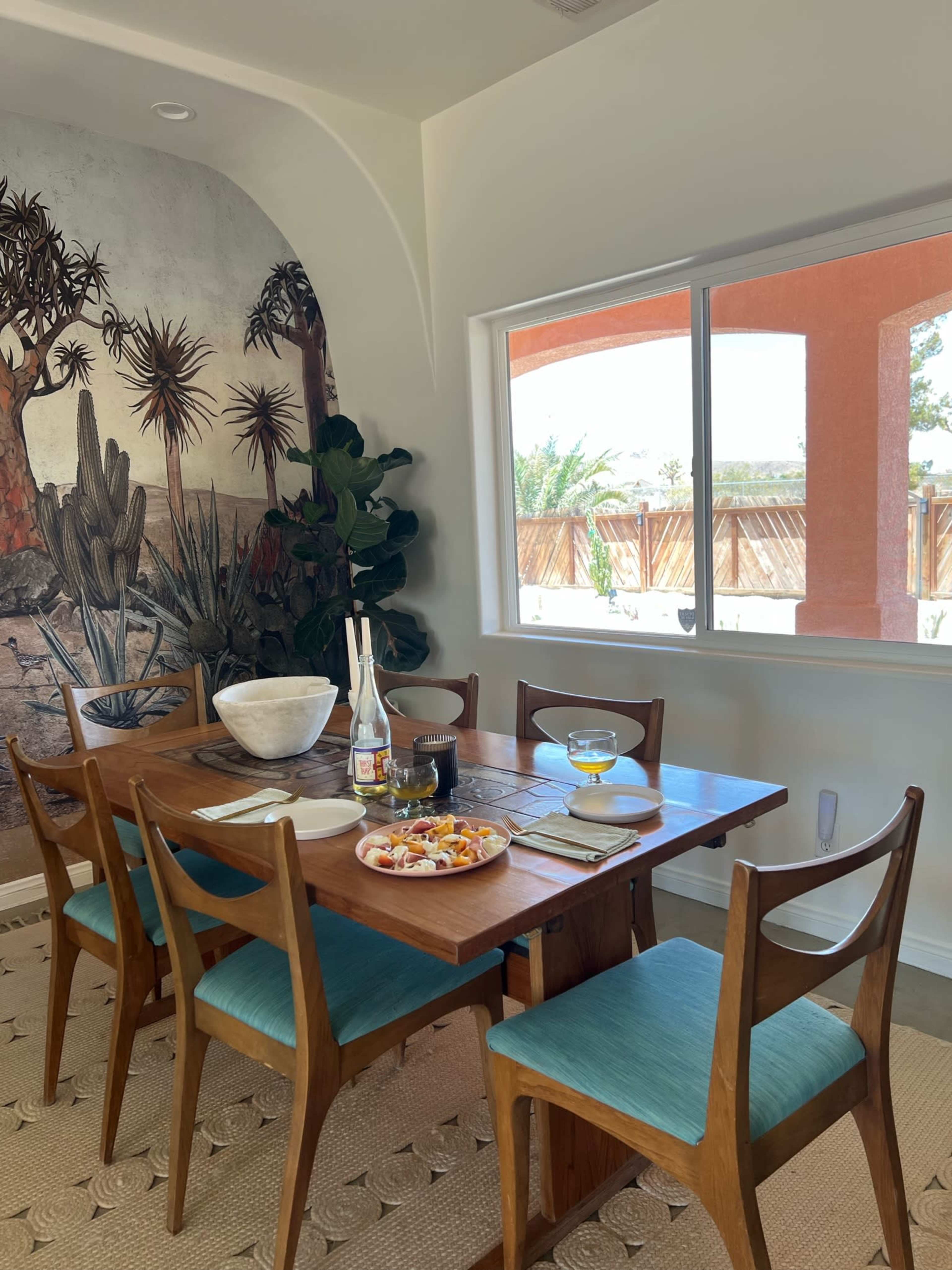 A dining table set with dishes and drinks is positioned near a large window, showcasing a mural of a desert landscape and a green plant in the corner.