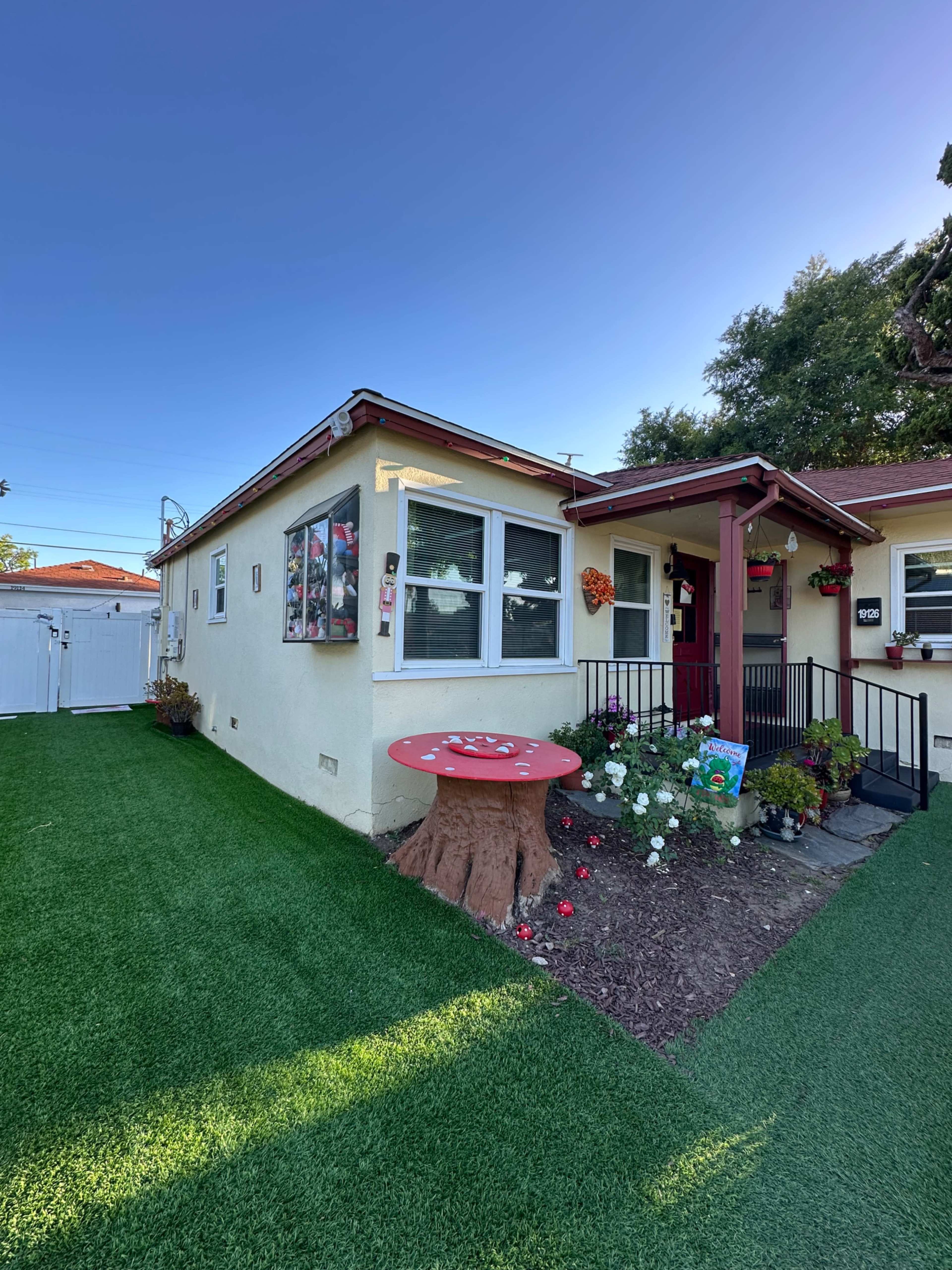 The image shows a small, single-story house with a landscaped front yard featuring artificial grass, a round red table made to look like a tree stump, and flower beds near the entrance.