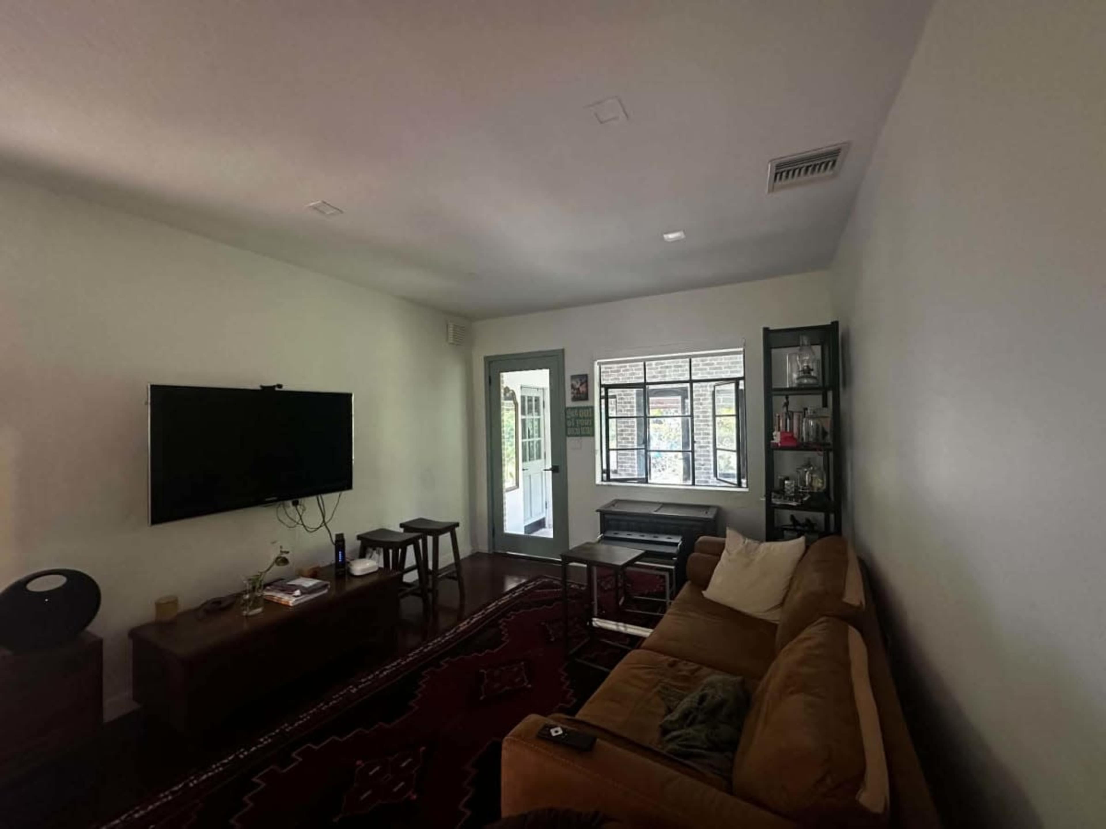 The image shows a living room with a television mounted on the wall, a brown sofa, a piano near a set of large windows, and various furniture pieces including a coffee table.