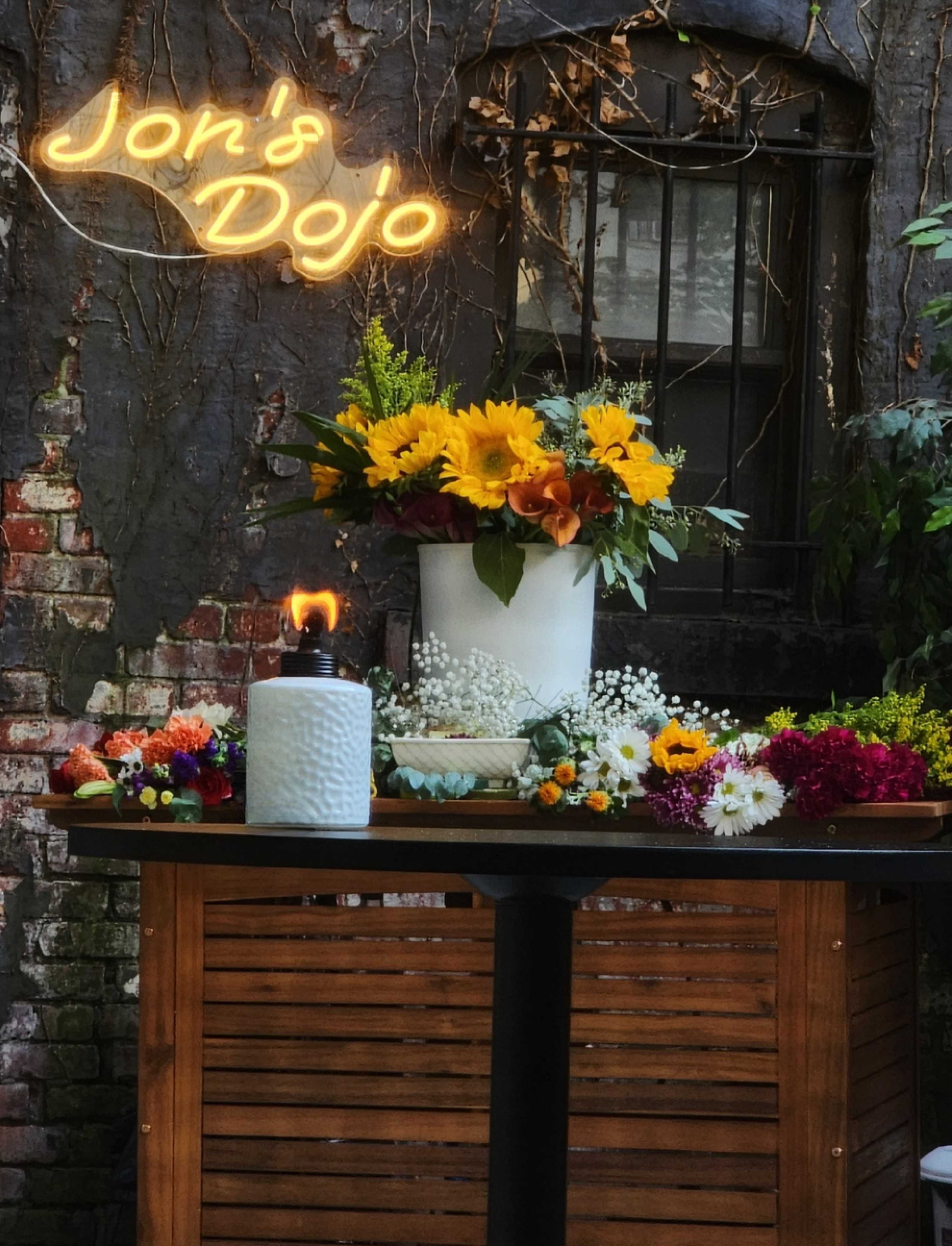 A wooden table adorned with colorful flowers and a candle sits beneath a neon sign that reads "Jon's Dojo" against a brick wall covered in vines.