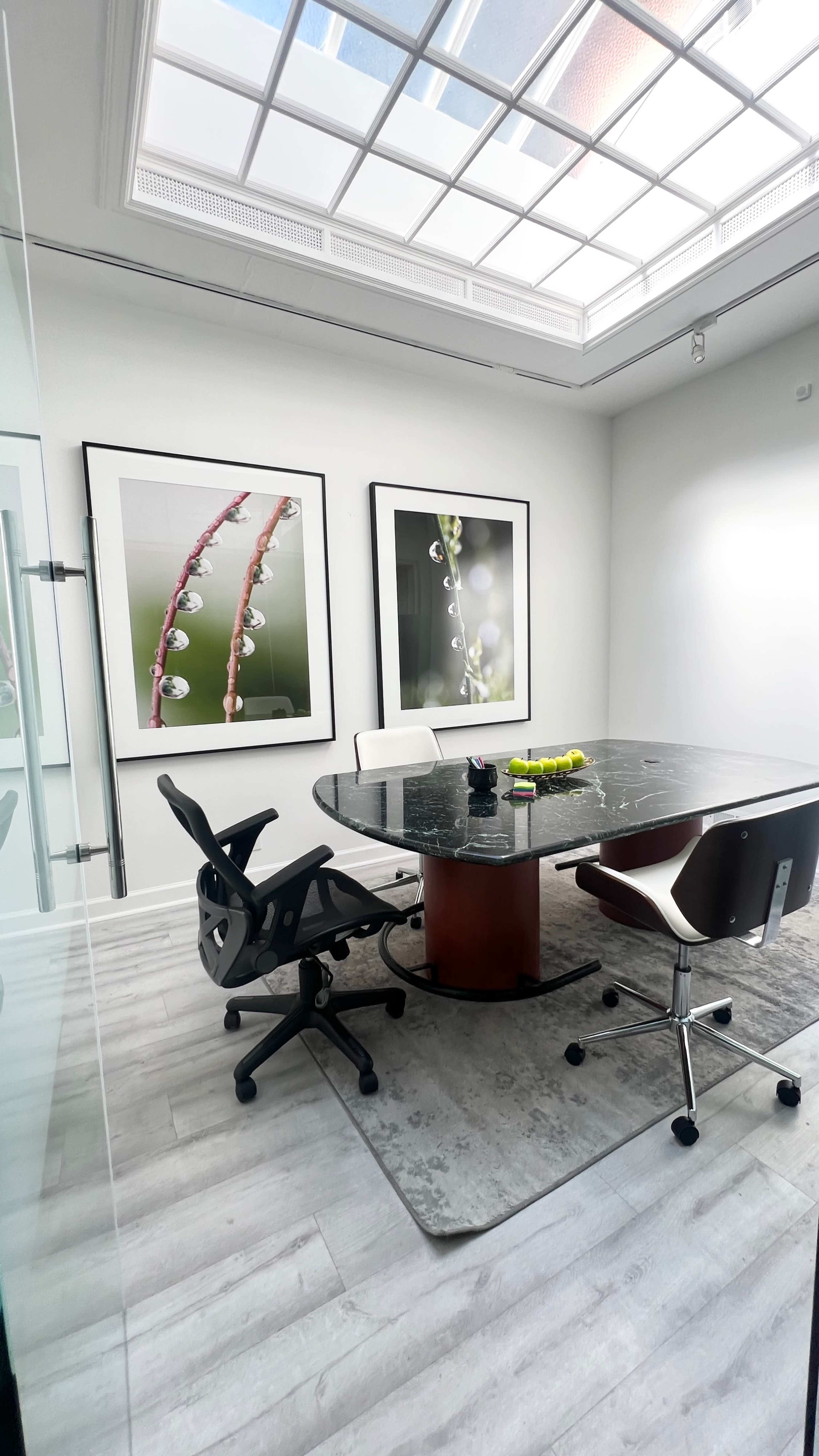 A conference room features a round table with black marble and two wheeled chairs, accented by large framed photographs of water droplets on plants.