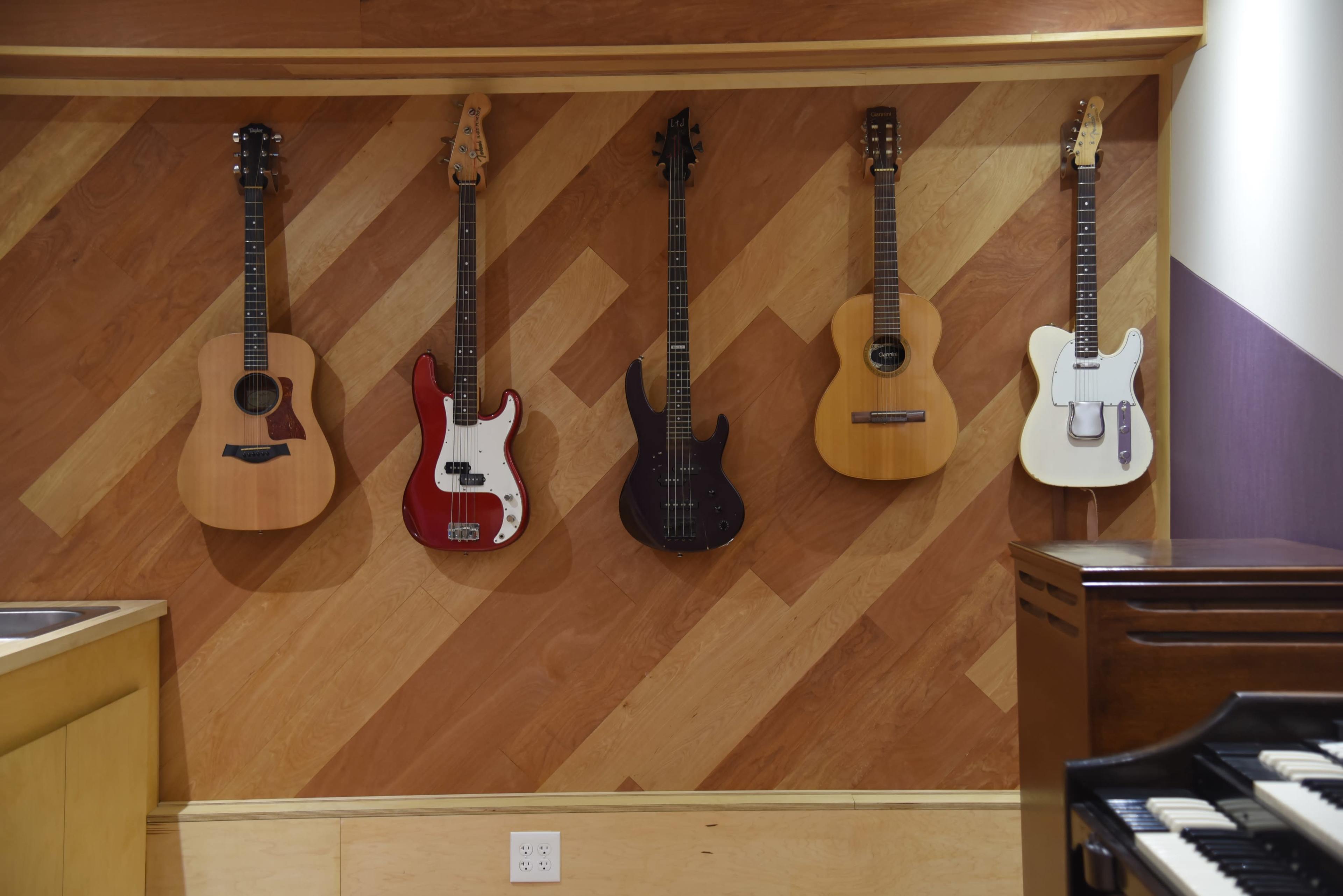 A row of five guitars of varying types and colors is displayed against a wood-paneled wall.