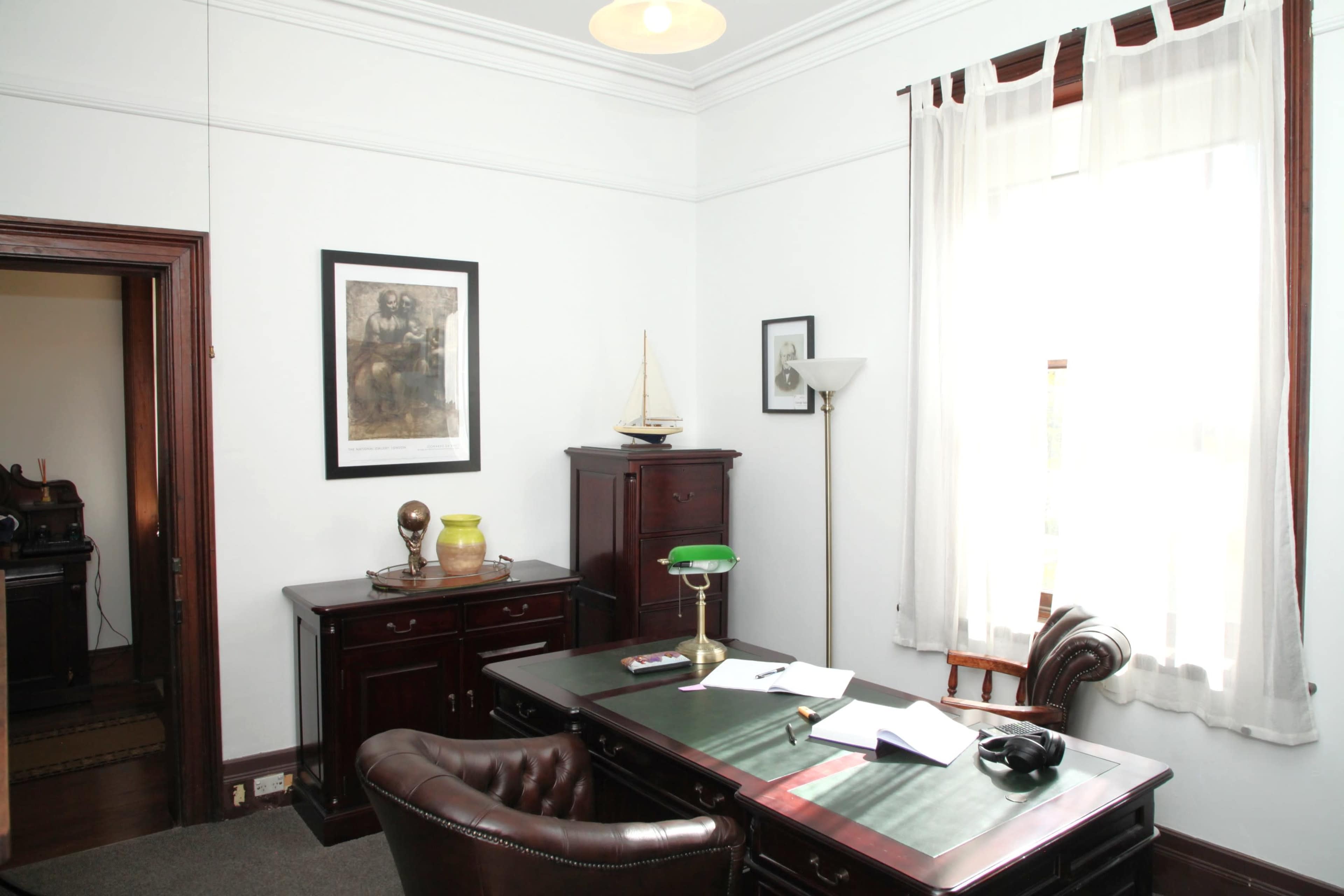 The image shows a well-organized office with a wooden desk, a chair, a lamp, and several framed artworks on the walls.