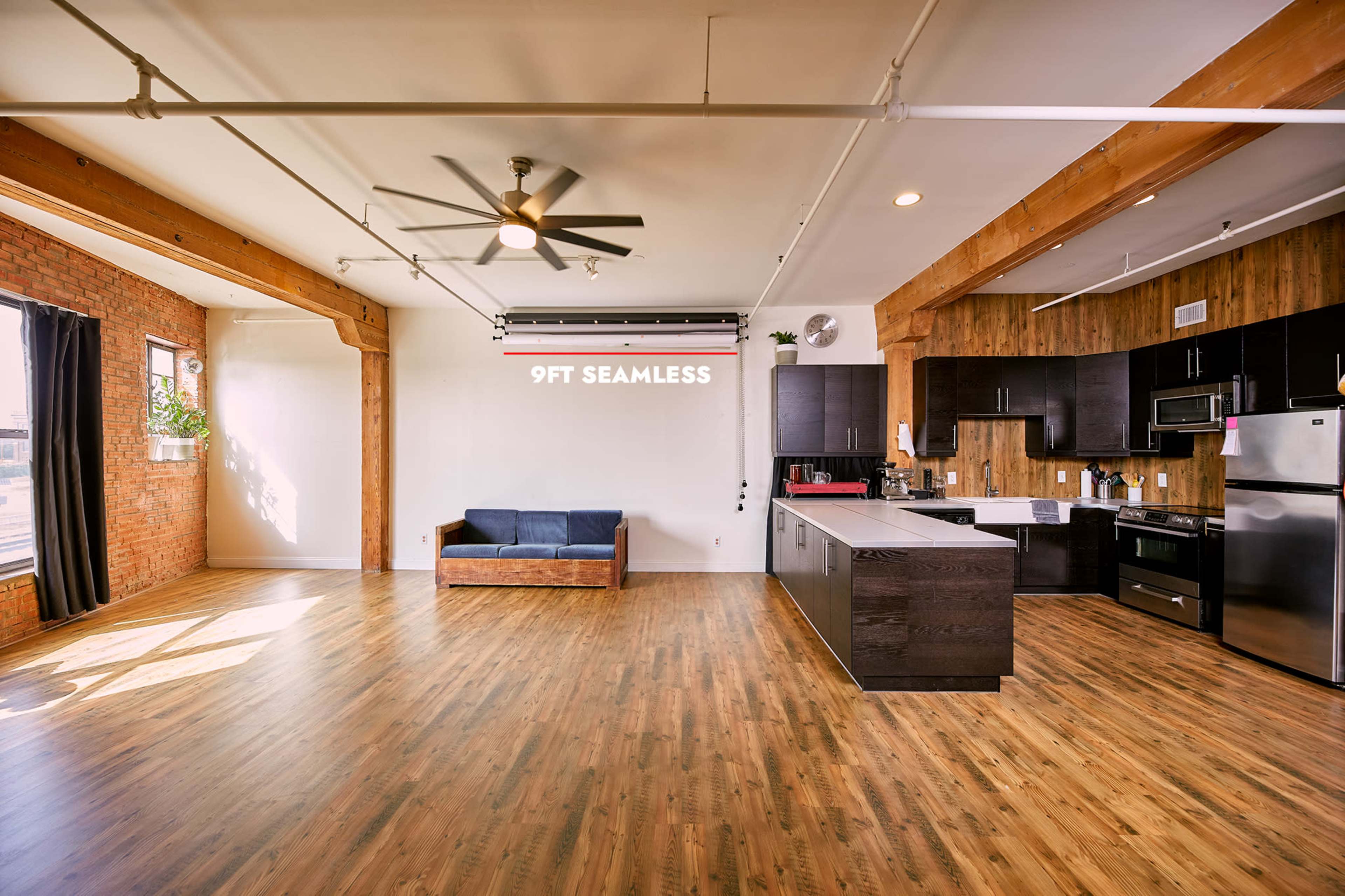 An open living space features a kitchen with dark cabinets, a wooden accent wall, a blue couch, and large windows allowing natural light to fill the room.