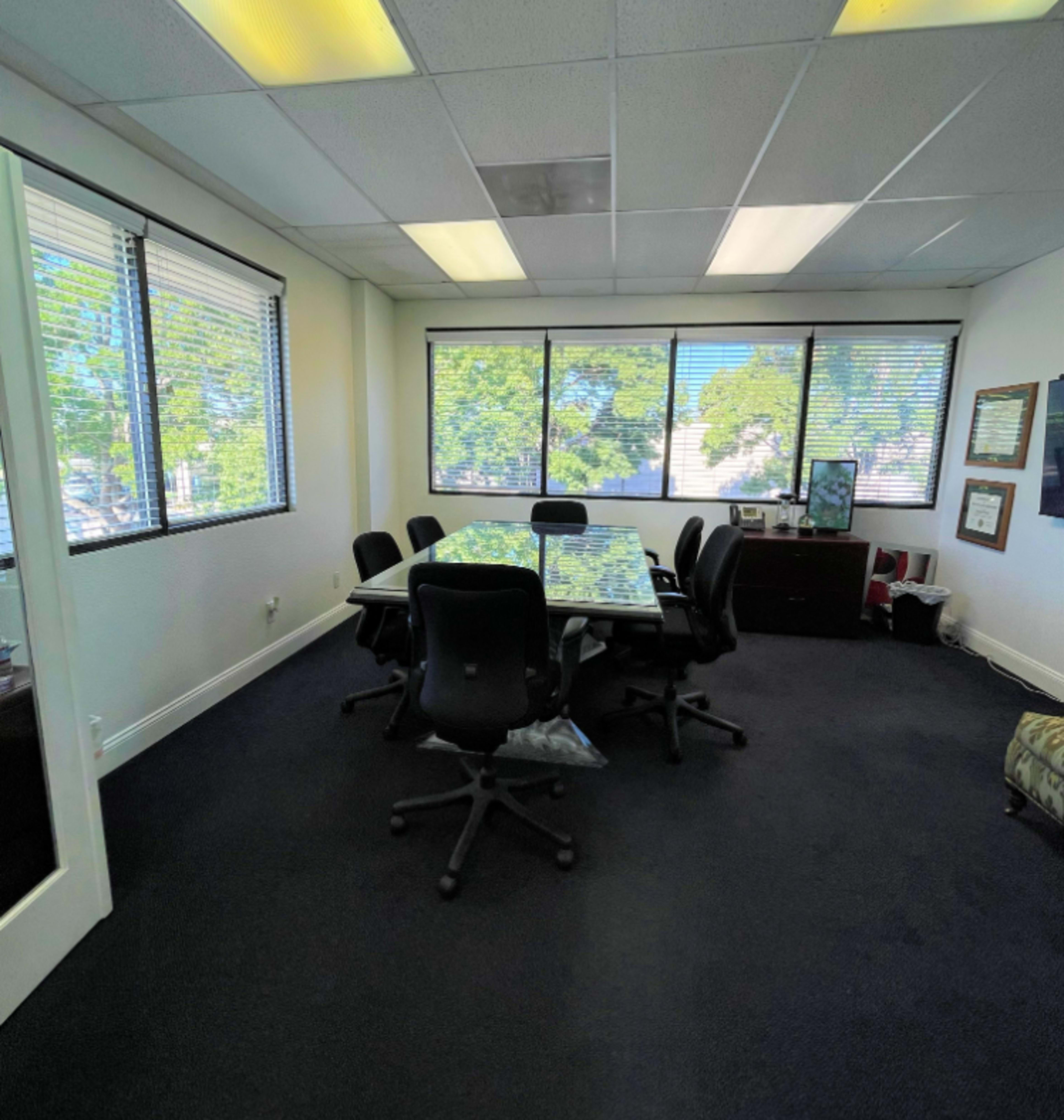 A conference room with a large glass table surrounded by black chairs and windows providing natural light.