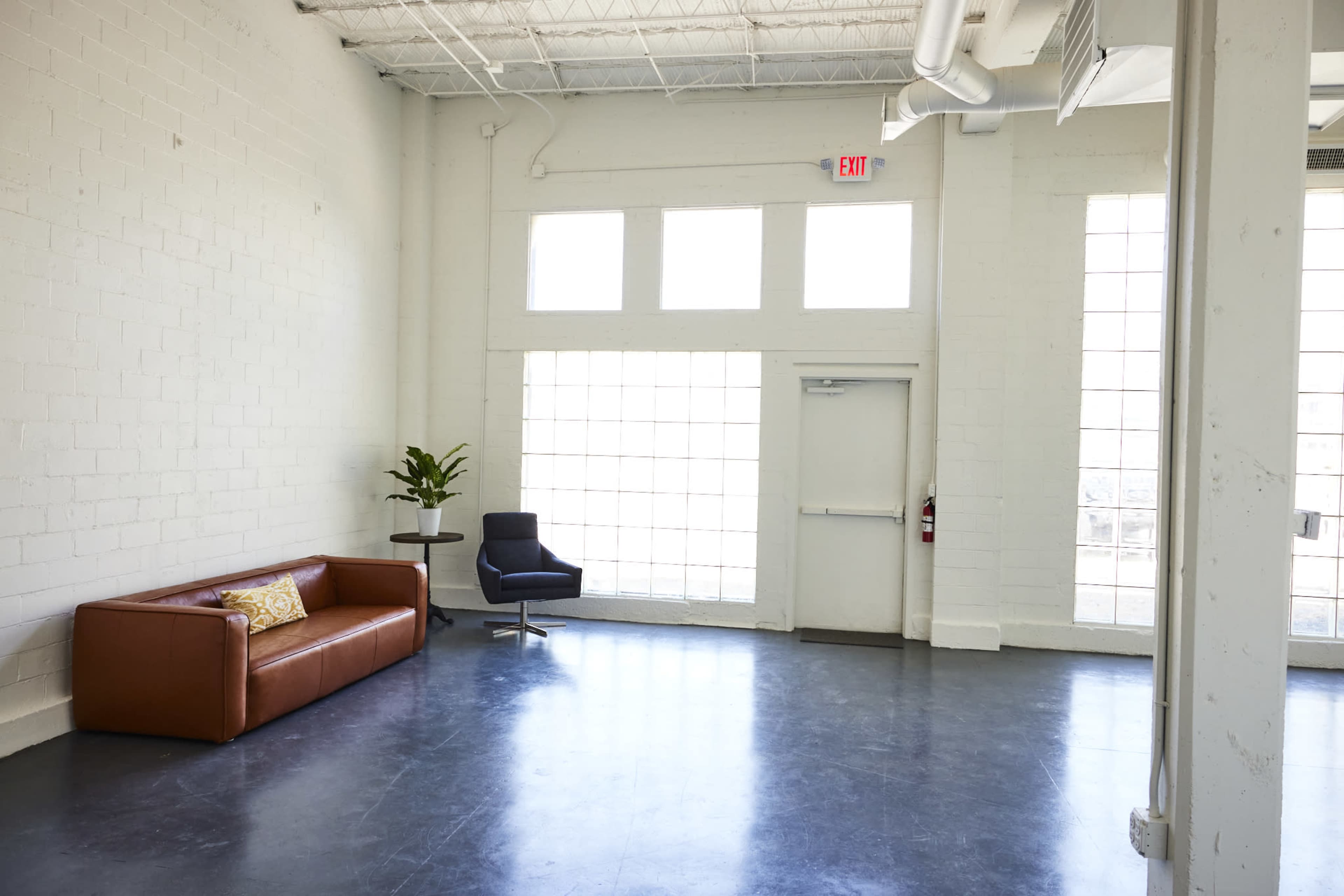 The image shows a spacious, empty room with large windows, a brown leather sofa, a modern chair, and a potted plant against a white wall.