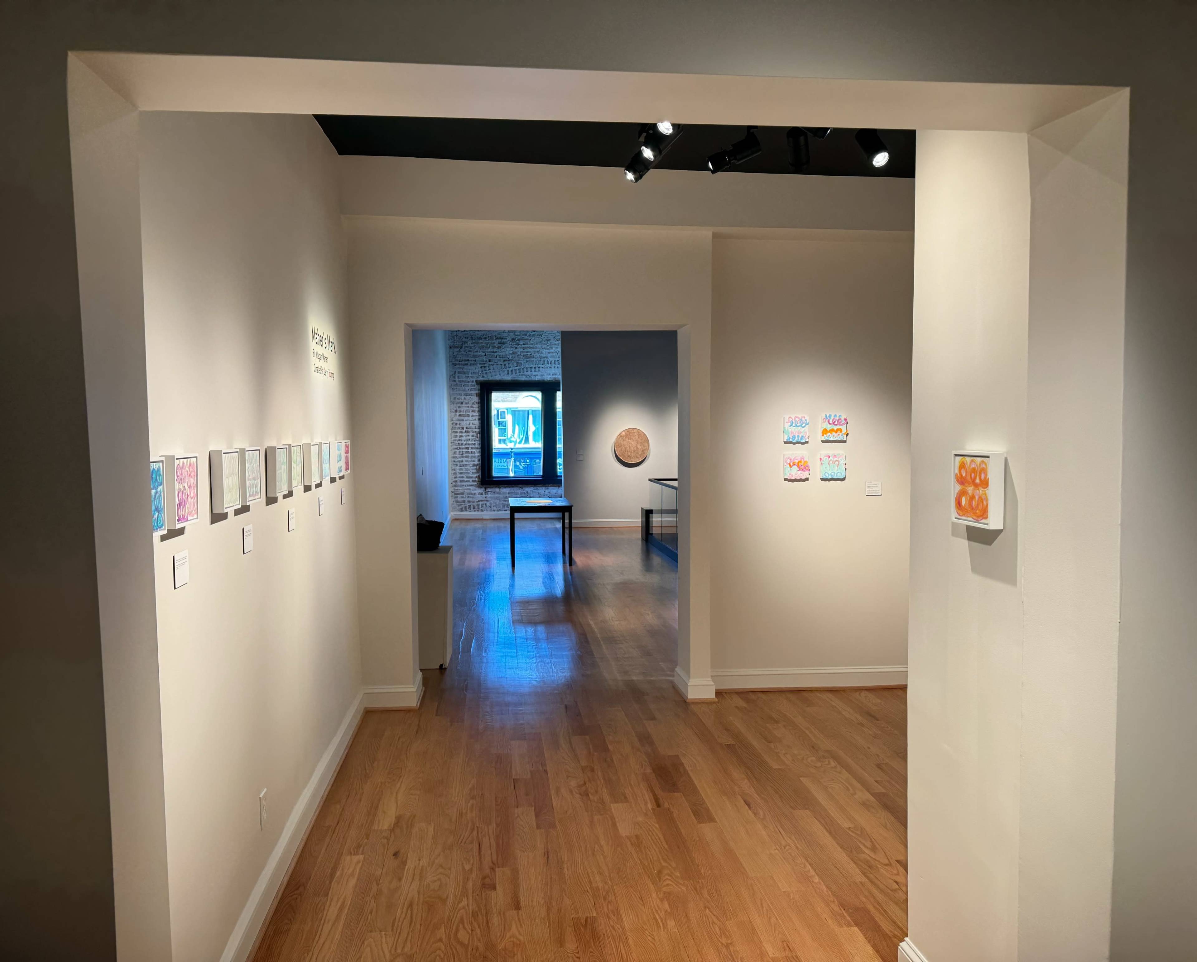 A gallery interior with white walls, wooden flooring, and artwork displayed on the walls and tables.