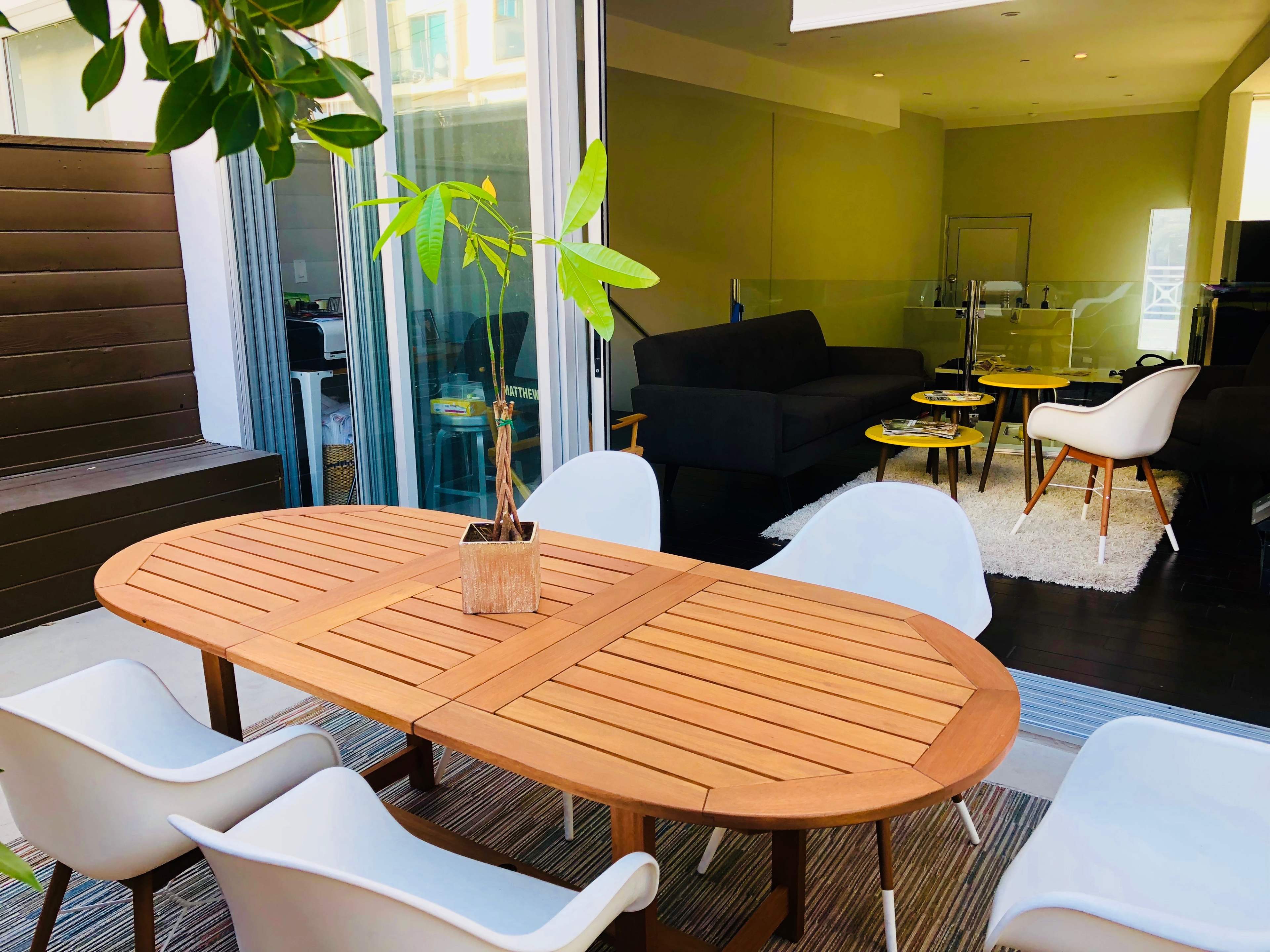 A wooden oval dining table with a potted plant in the center is surrounded by white chairs on a patio, leading into a brightly lit indoor space with a couch and two small tables.