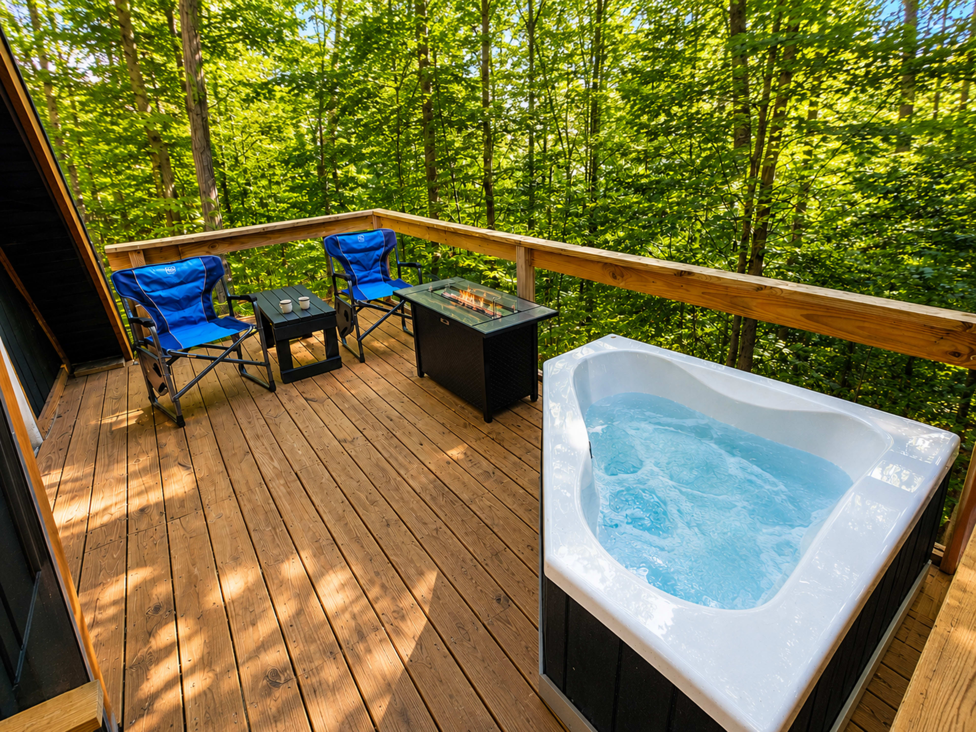 A wooden deck surrounded by trees features a hot tub, two blue chairs, and a small table with a fire pit.