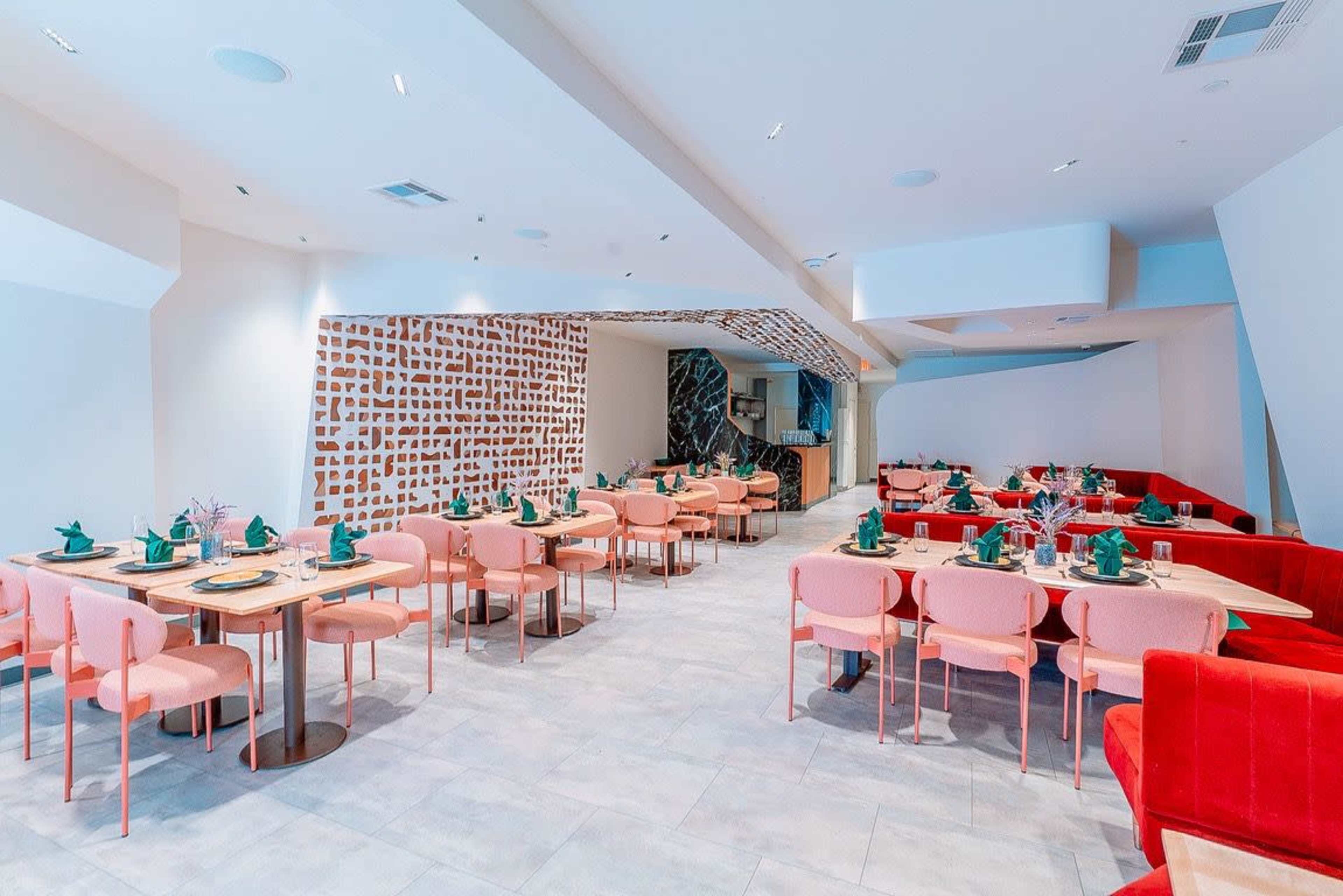 The image shows a modern restaurant interior with pink chairs, wooden tables, and green table settings against a light-colored wall with decorative patterns.