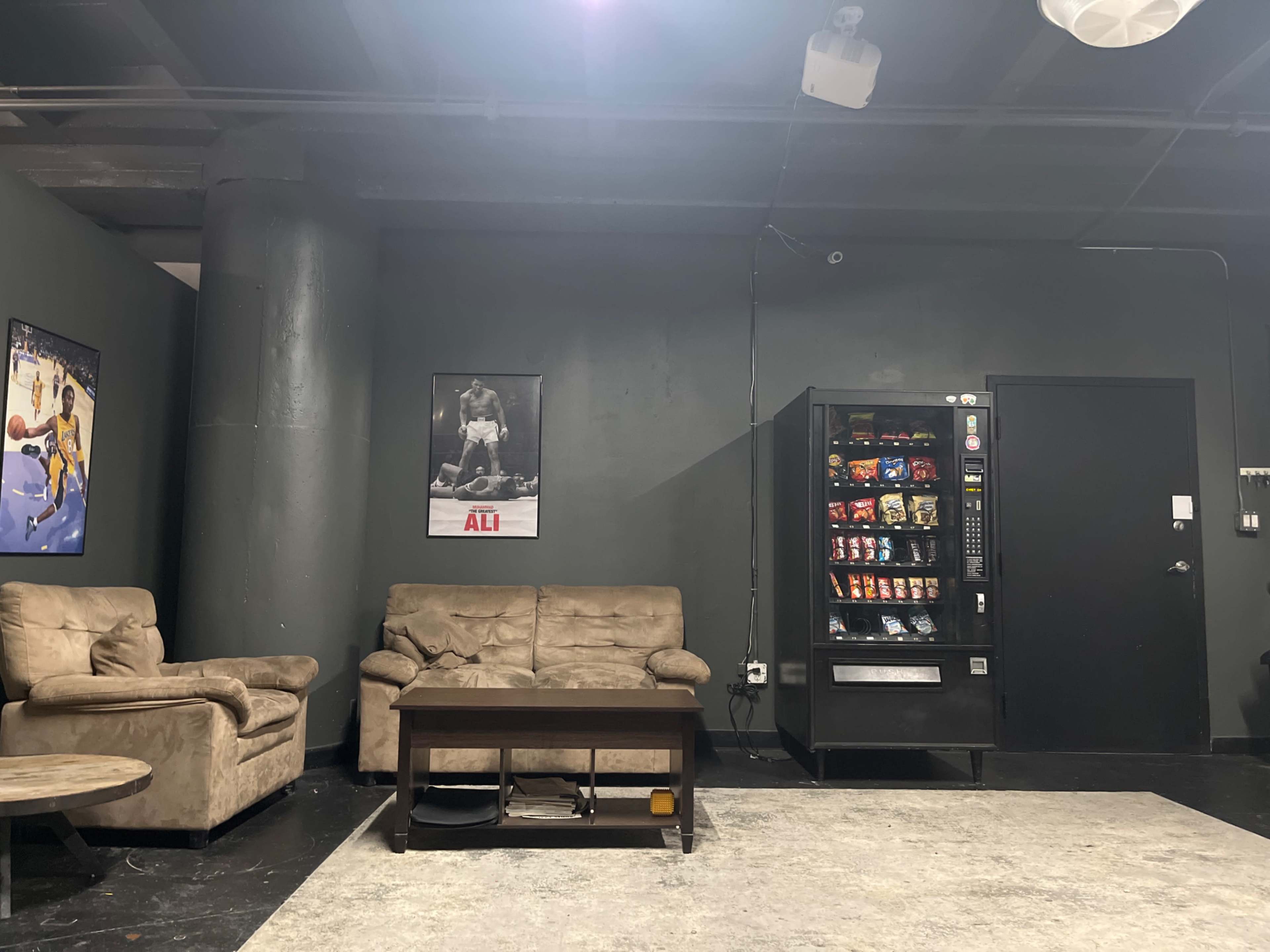 A dimly lit lounge area with two beige couches, a wooden coffee table, a vending machine, and a poster of a boxer on the wall.