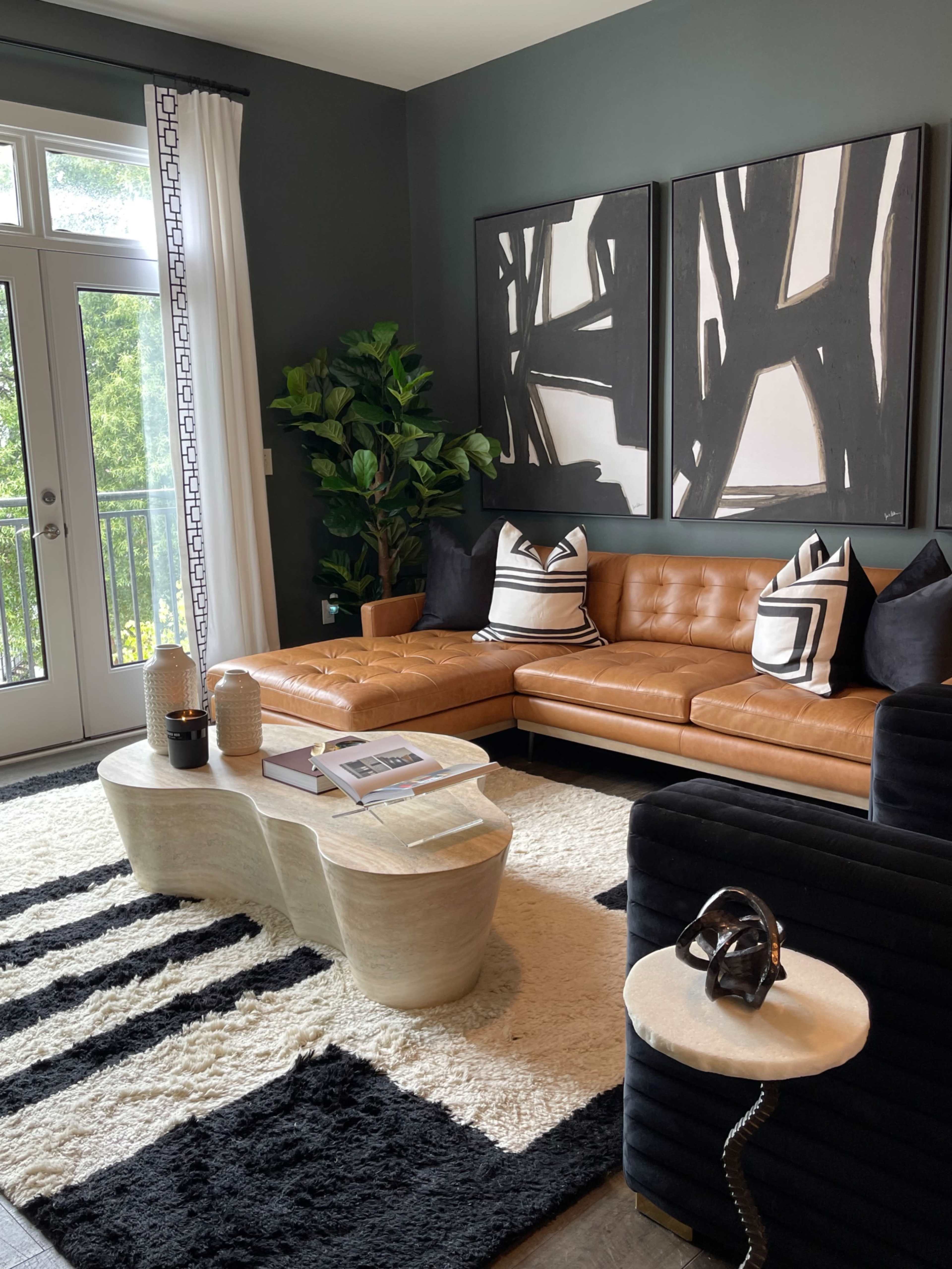 The living room features a brown leather sectional sofa, a light-colored abstract coffee table, a black and white area rug, and two large artworks on the wall.