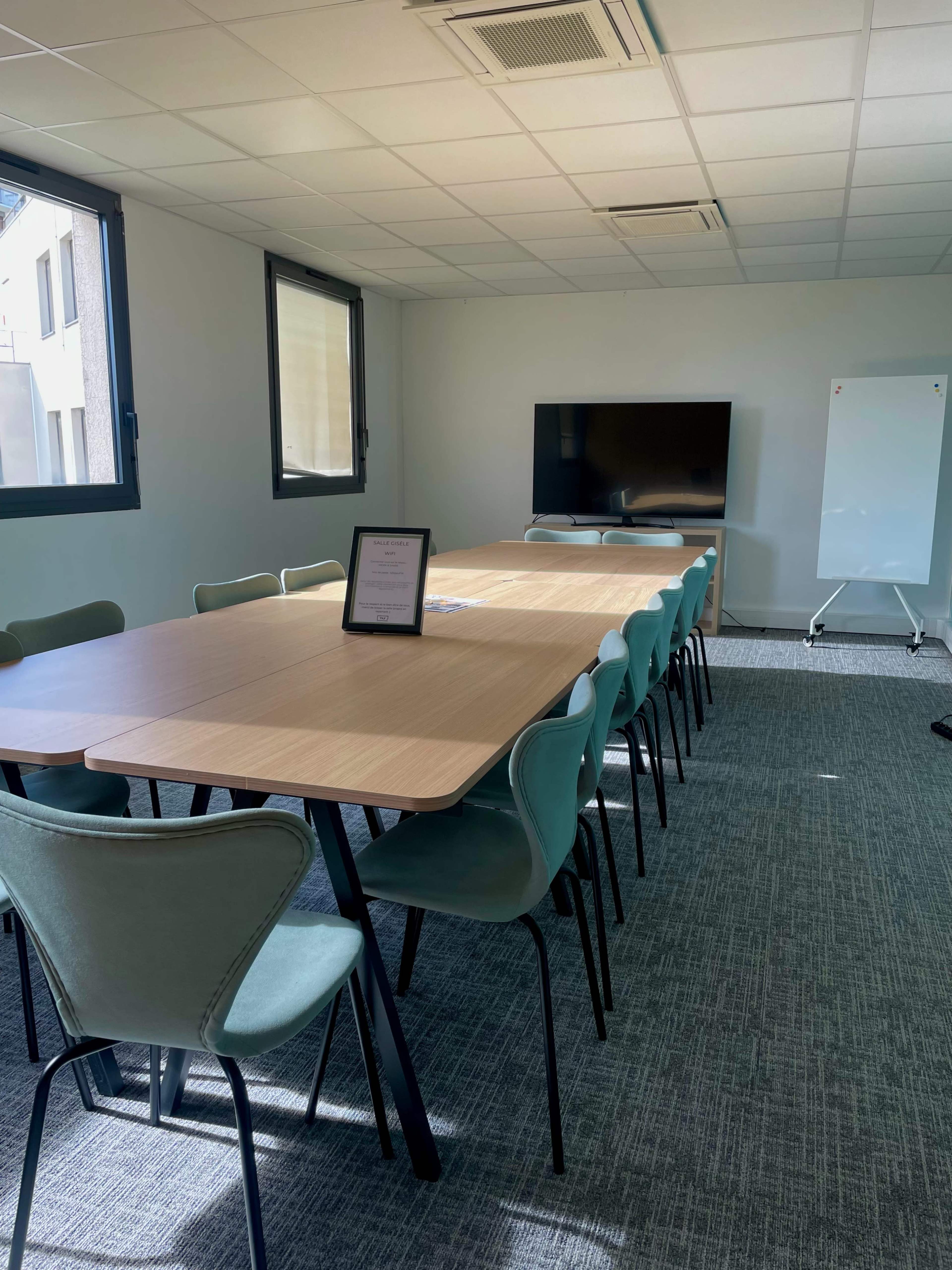 A modern meeting room features a long wooden table surrounded by green chairs, with a large TV screen and a whiteboard along the wall.