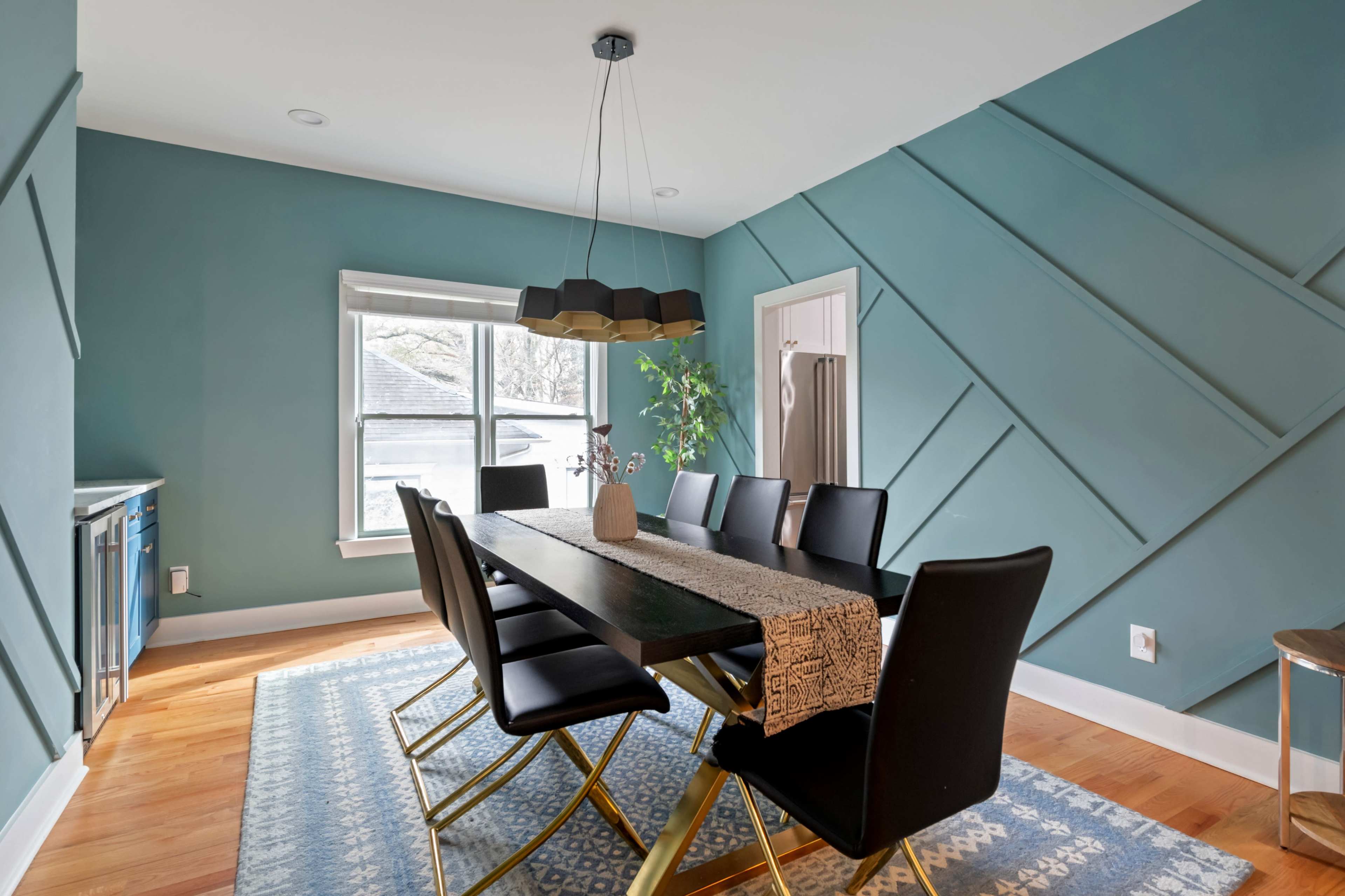 The image shows a modern dining room with a wooden table, black chairs, a large window, and teal walls featuring geometric paneling.