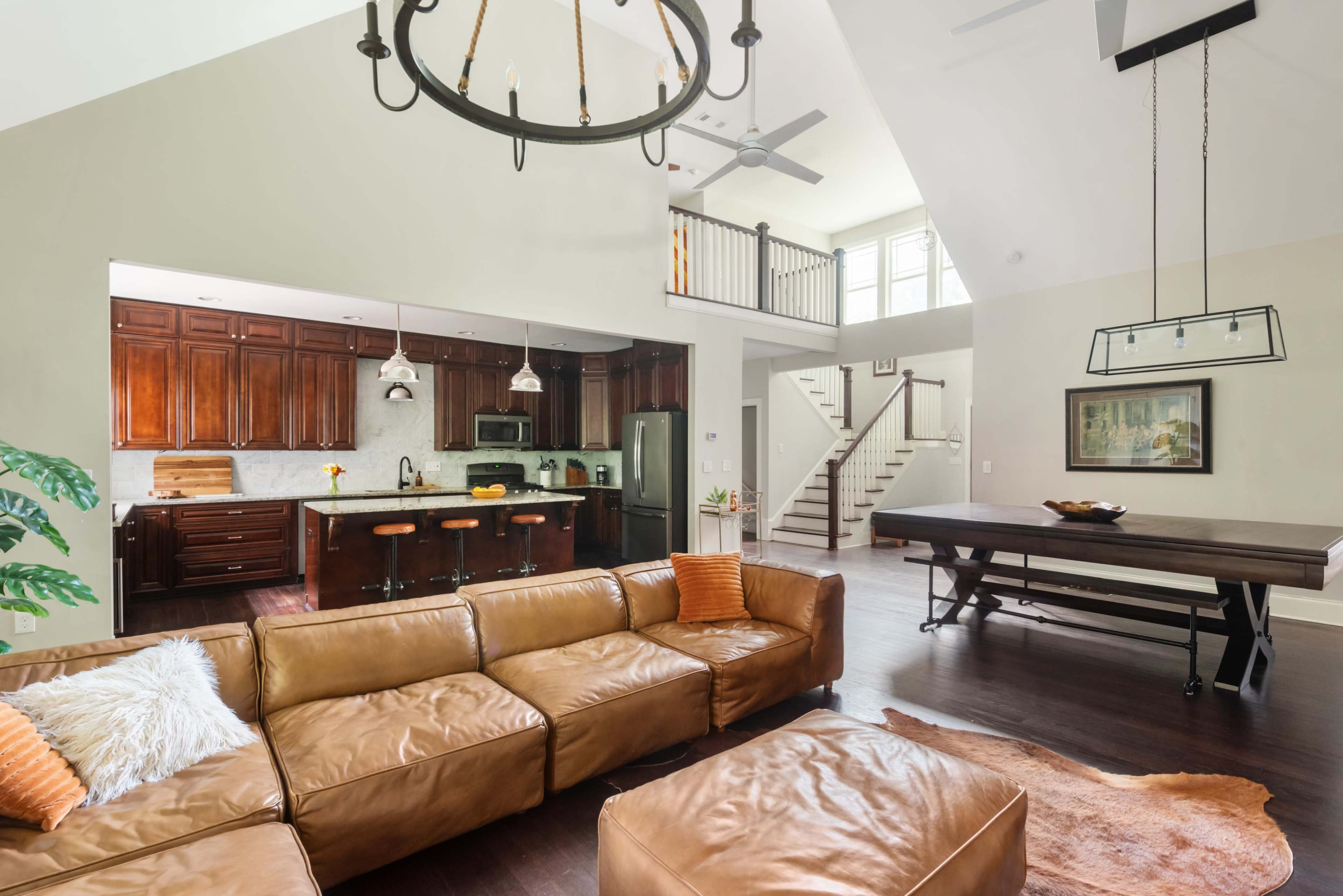 The image shows a spacious living area with a leather sectional sofa, a modern dining table, and a kitchen with dark wood cabinetry and pendant lights.