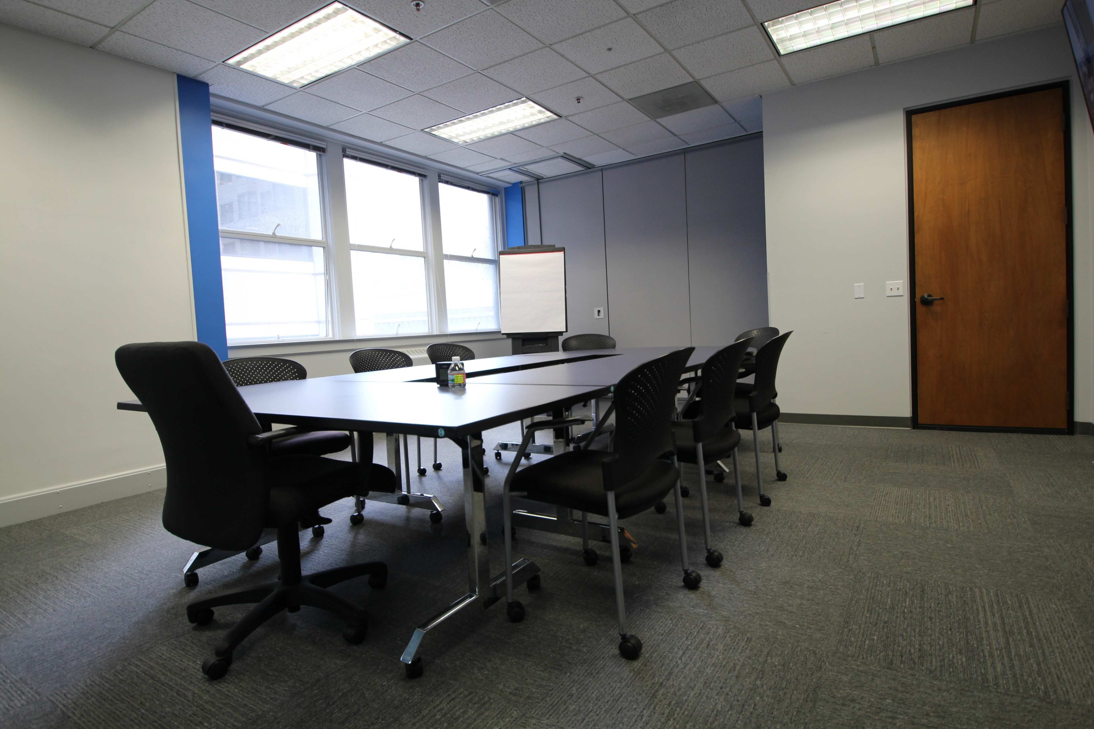 A conference room features a large table surrounded by eight chairs, with windows letting in natural light and a whiteboard mounted on the wall.