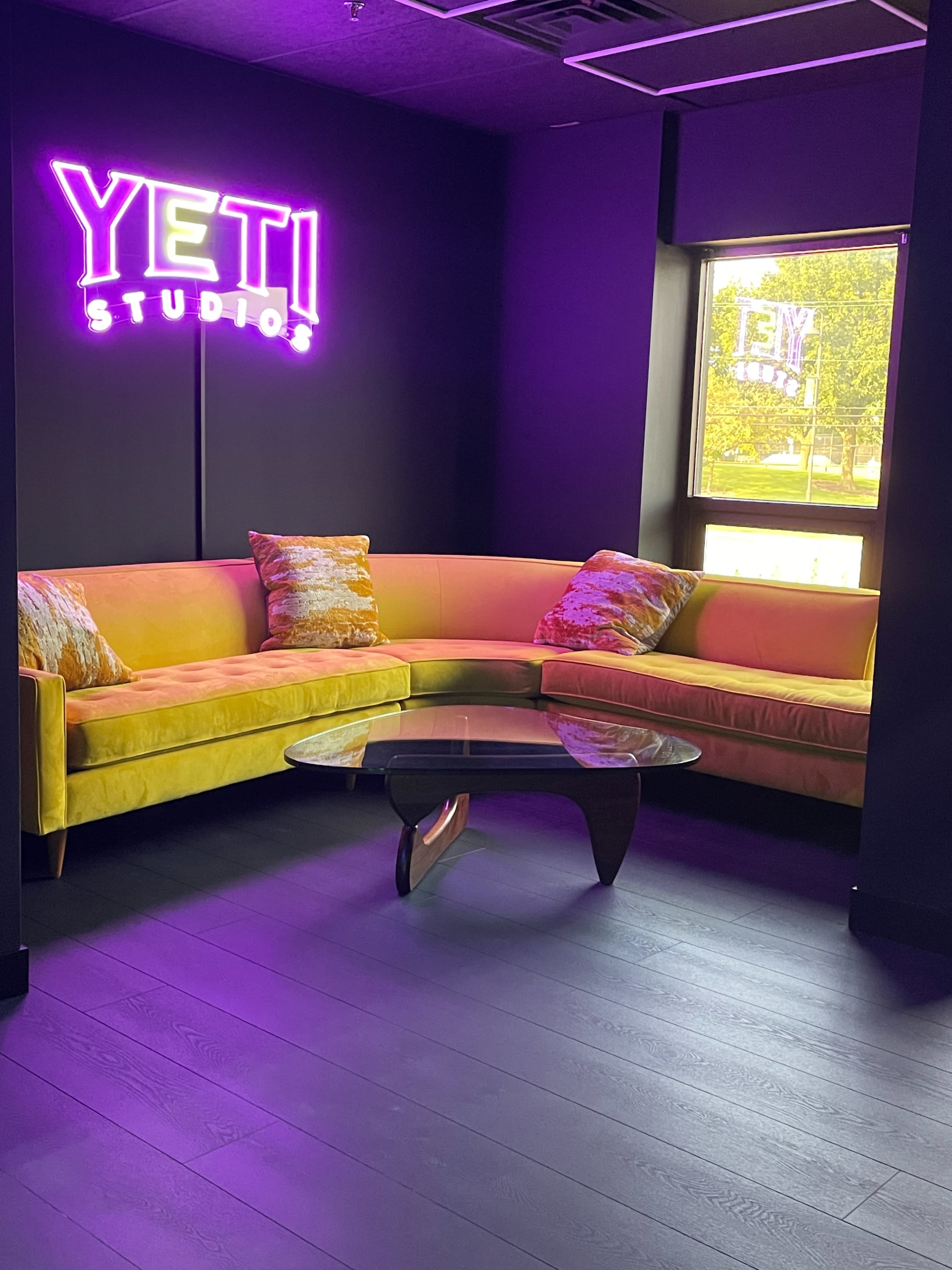 A bright yellow sectional sofa surrounds a glass coffee table in a modern studio lounge with purple walls and a neon sign reading "YETI STUDIOS."