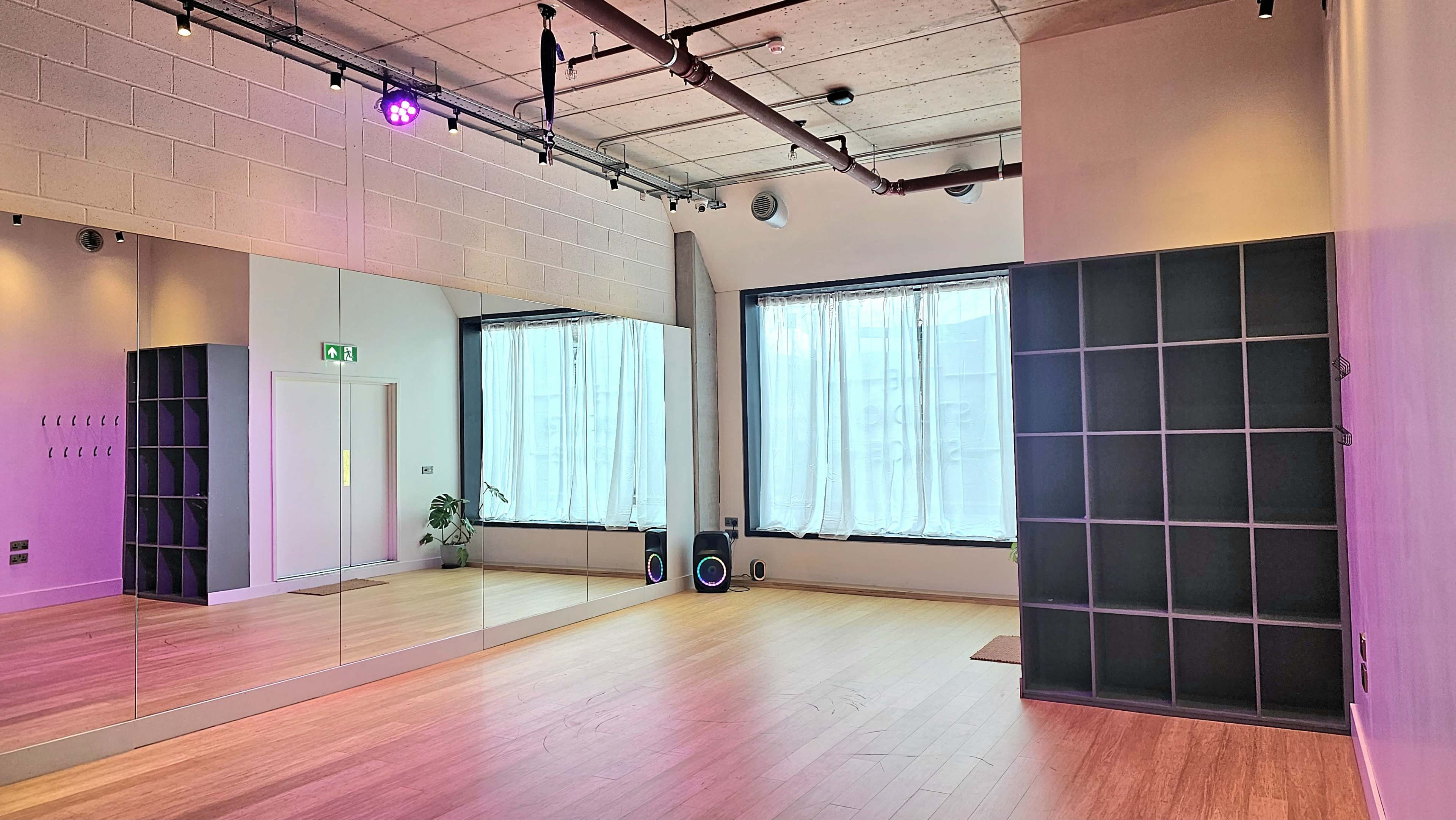 The image shows a light-filled studio with hardwood floors, large windows covered by sheer curtains, and a mirrored wall.