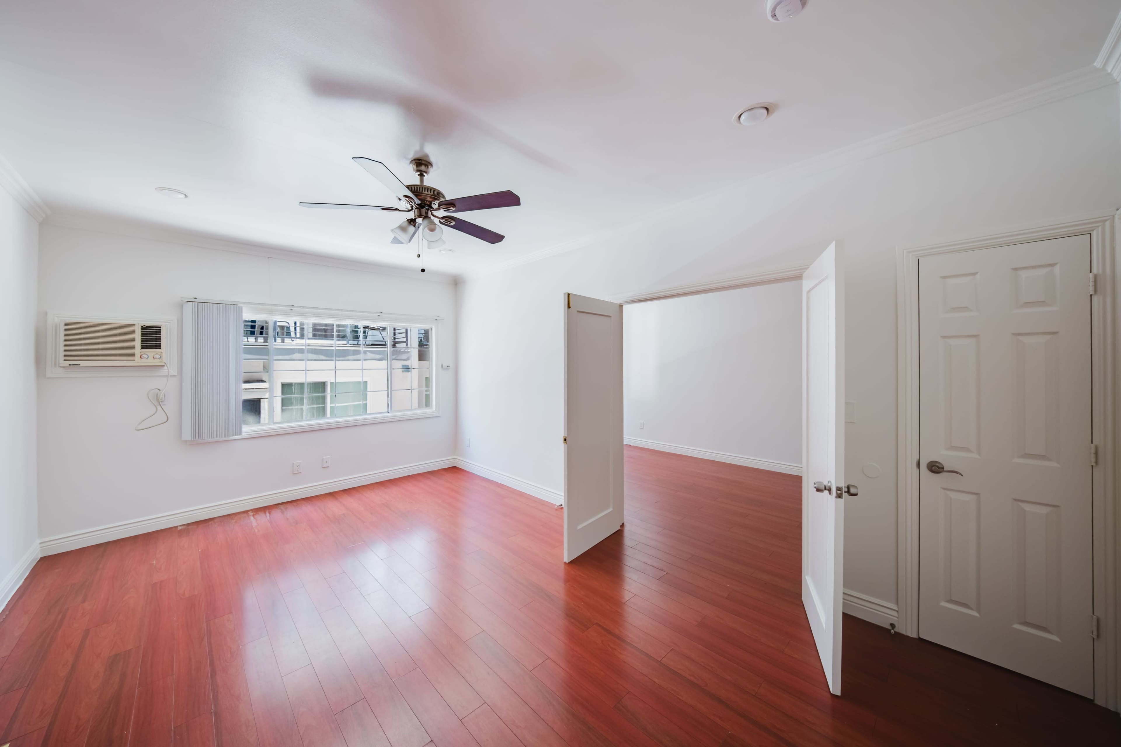 The image shows a sparsely furnished room with hardwood flooring, a ceiling fan, and a window allowing natural light.