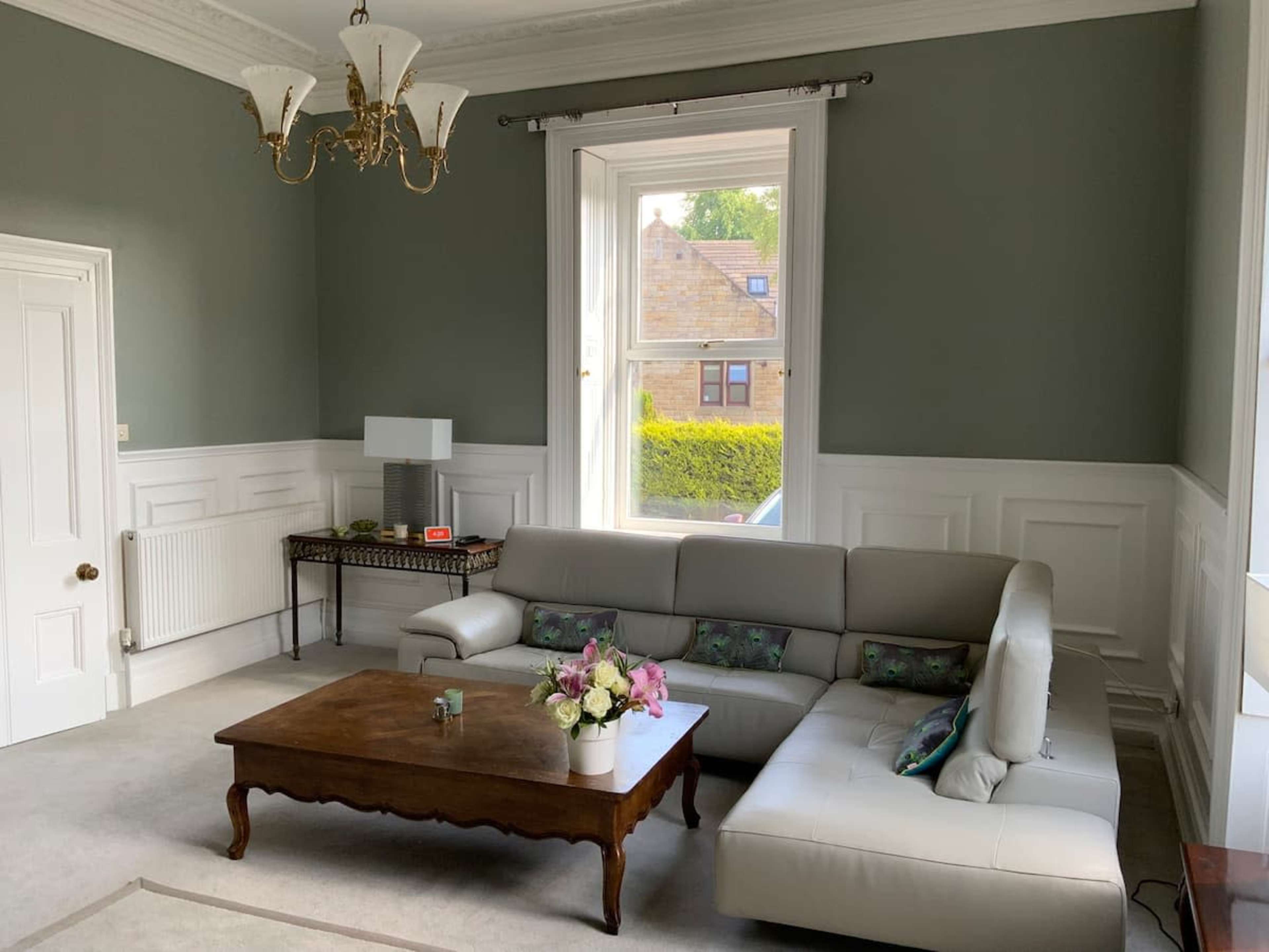 A modern living room features a light-colored sectional sofa, a wooden coffee table with flowers, and a window revealing a view of greenery outside.