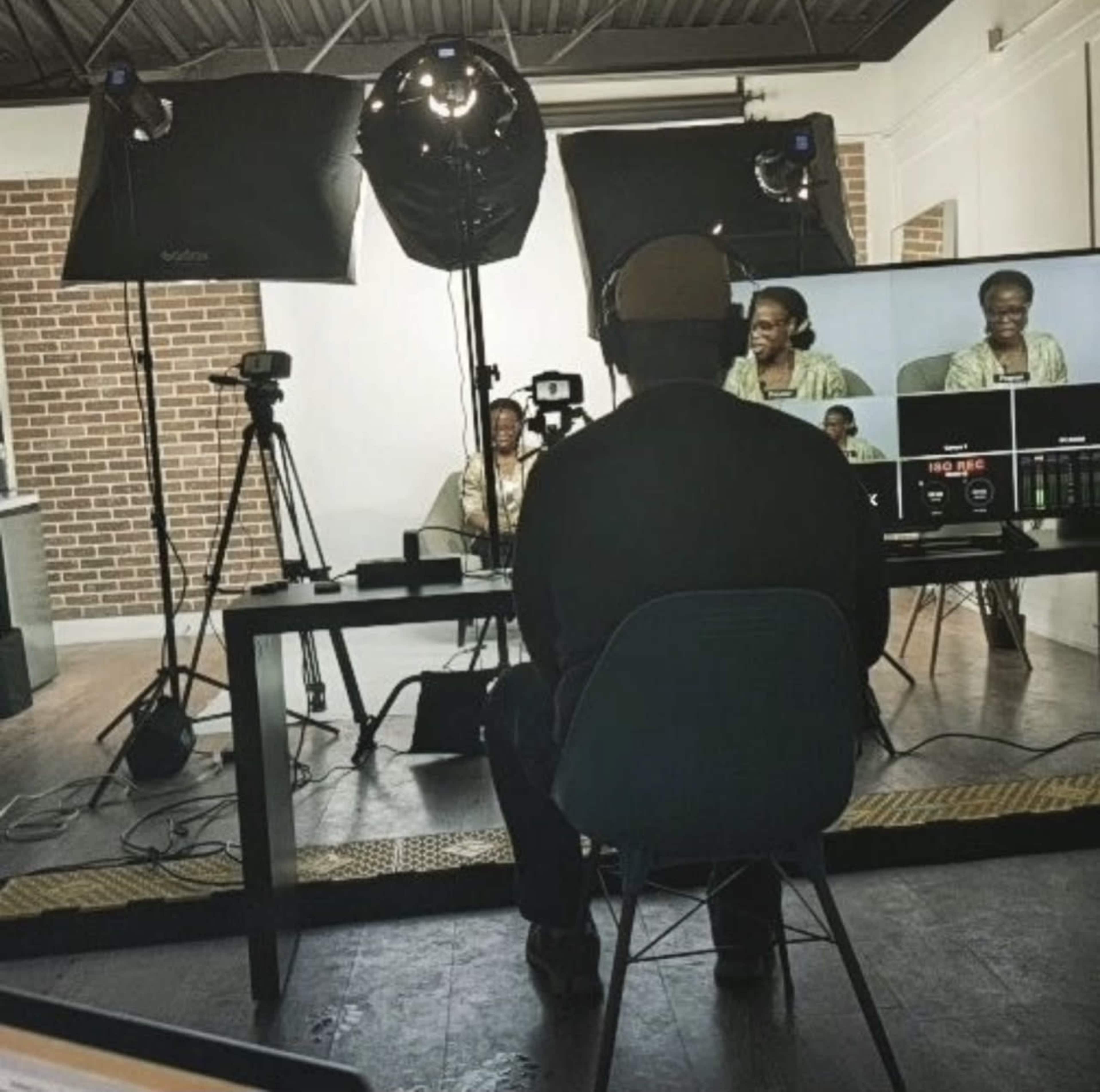 A person sits at a table facing multiple cameras and studio lights, while monitoring a live feed of a woman speaking in front of a brick wall.