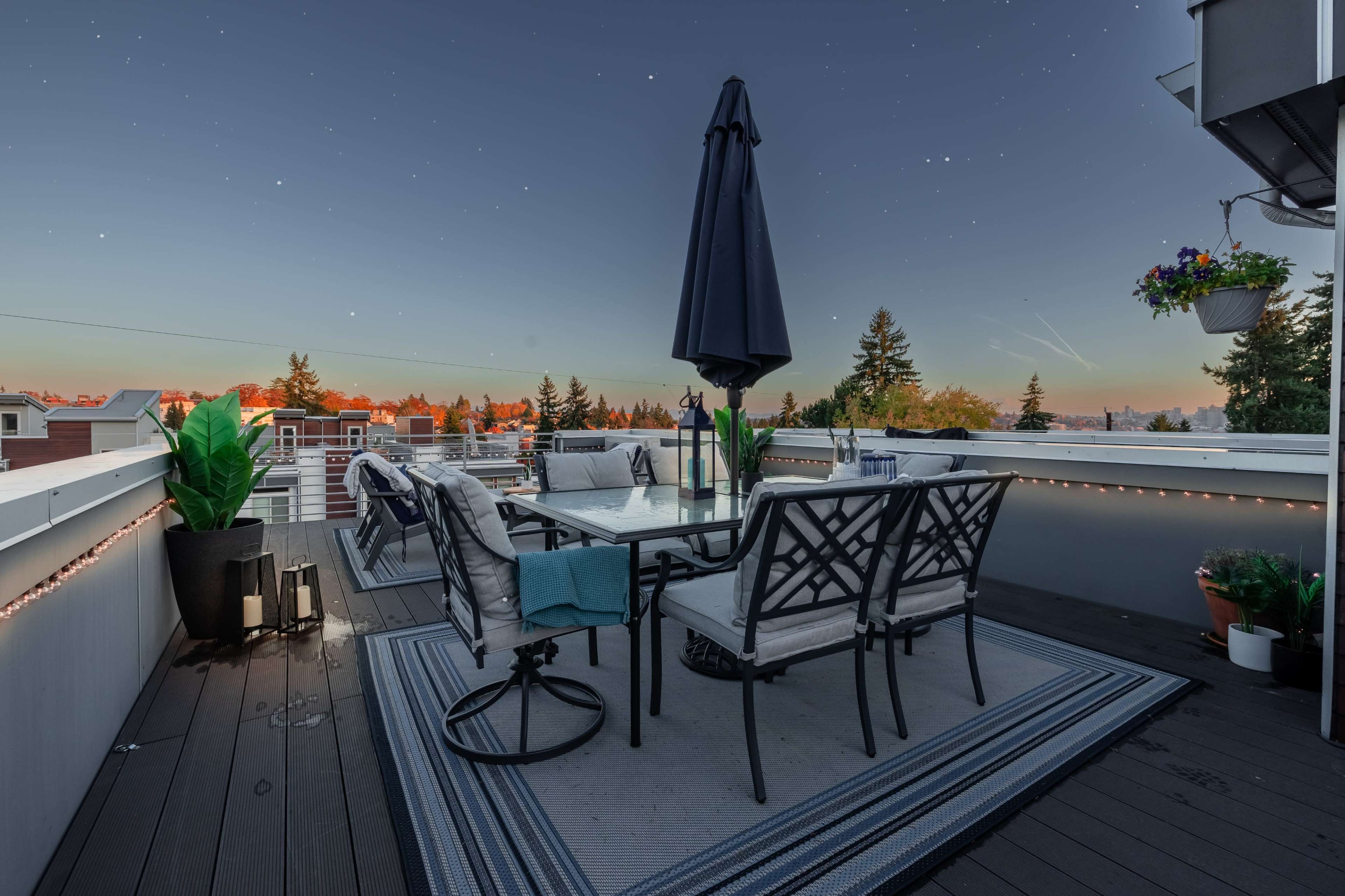 A rooftop terrace features a table surrounded by six chairs, with a large umbrella, overlooking a scenic view at dusk.