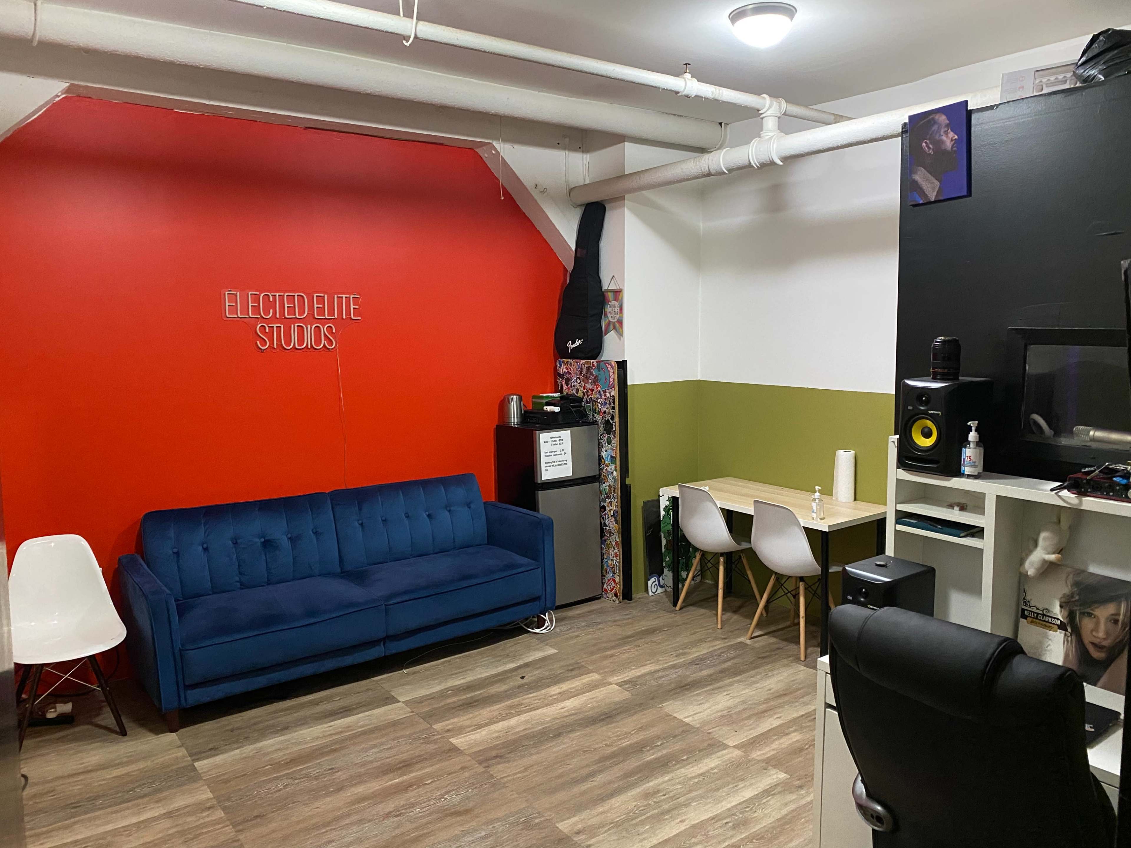 The image shows an interior studio with a red accent wall, a blue couch, a mini-fridge, a desk with equipment, and white chairs.