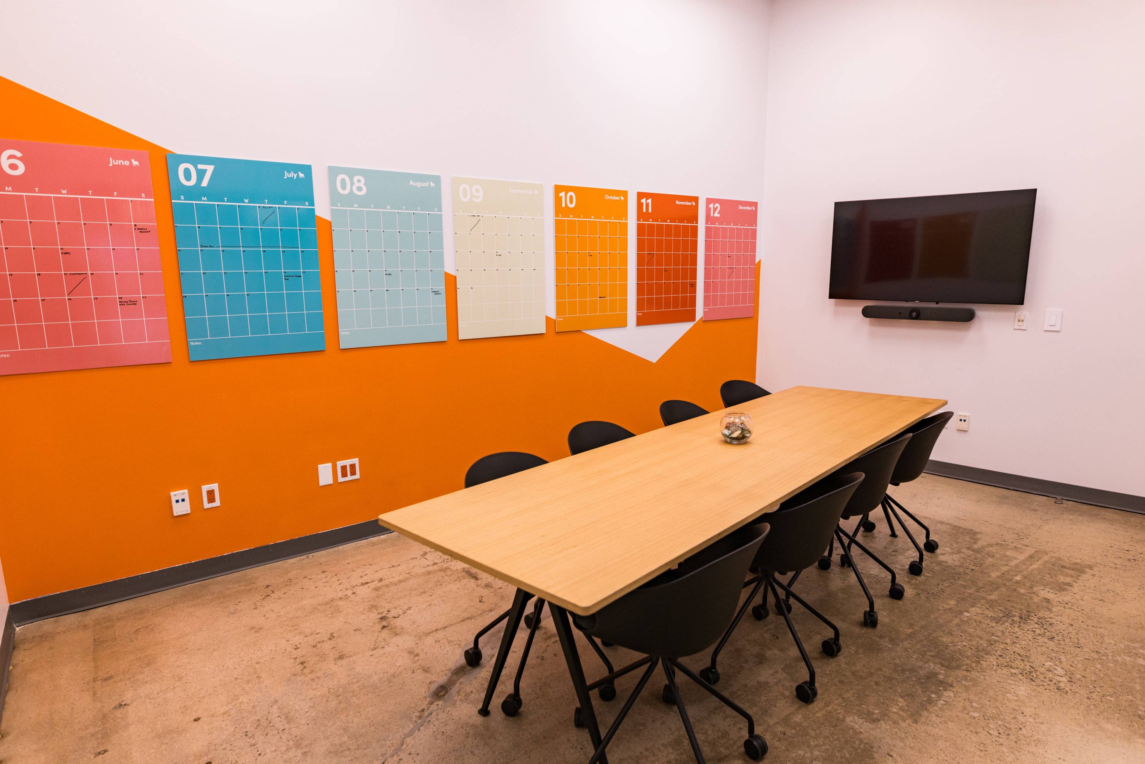 A meeting room features a long wooden table surrounded by black chairs, with colorful monthly calendars on the wall and a television mounted nearby.