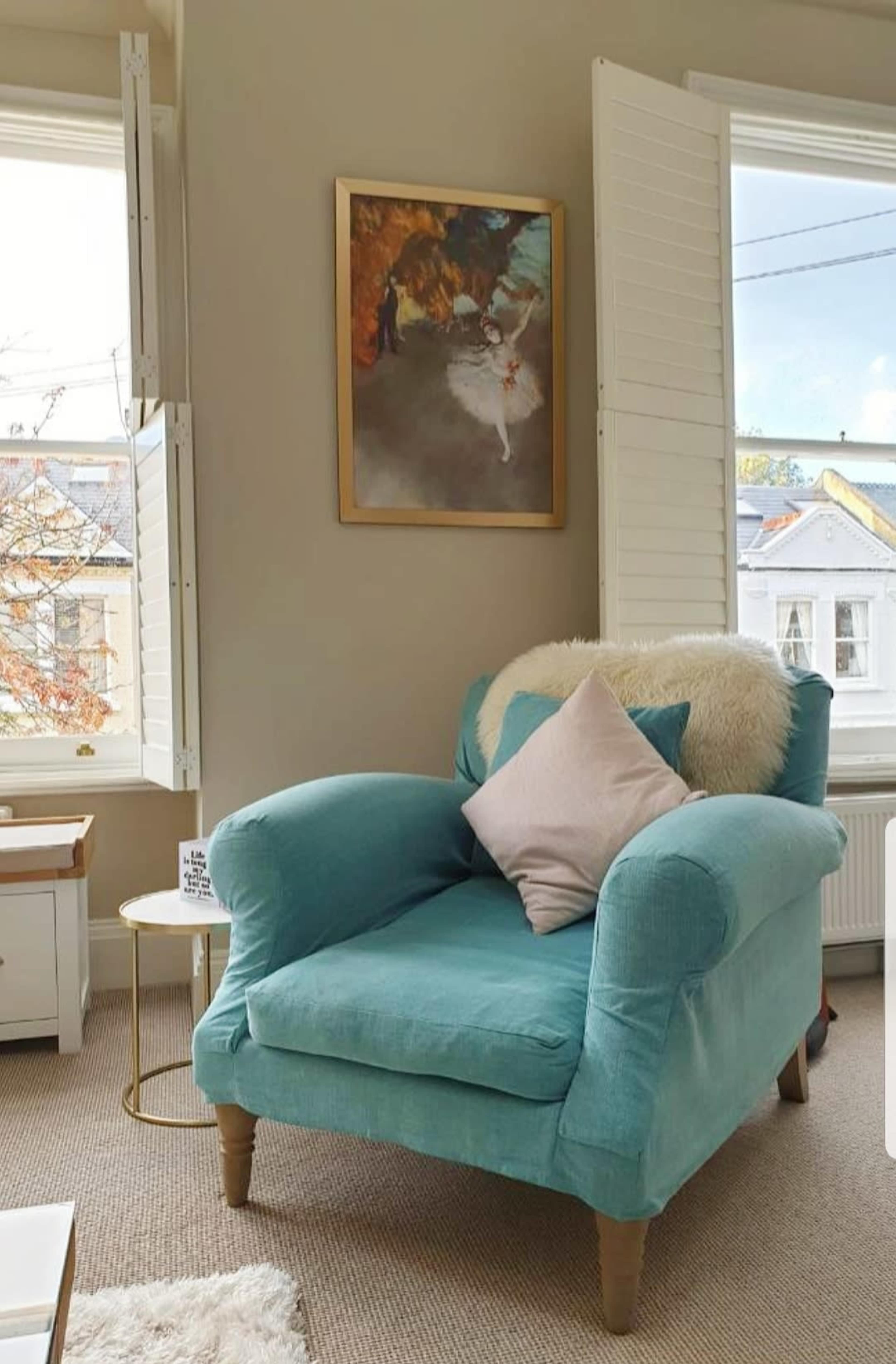 A light blue armchair with a pink pillow sits in a well-lit room, next to a small round table and a wall featuring a framed painting.