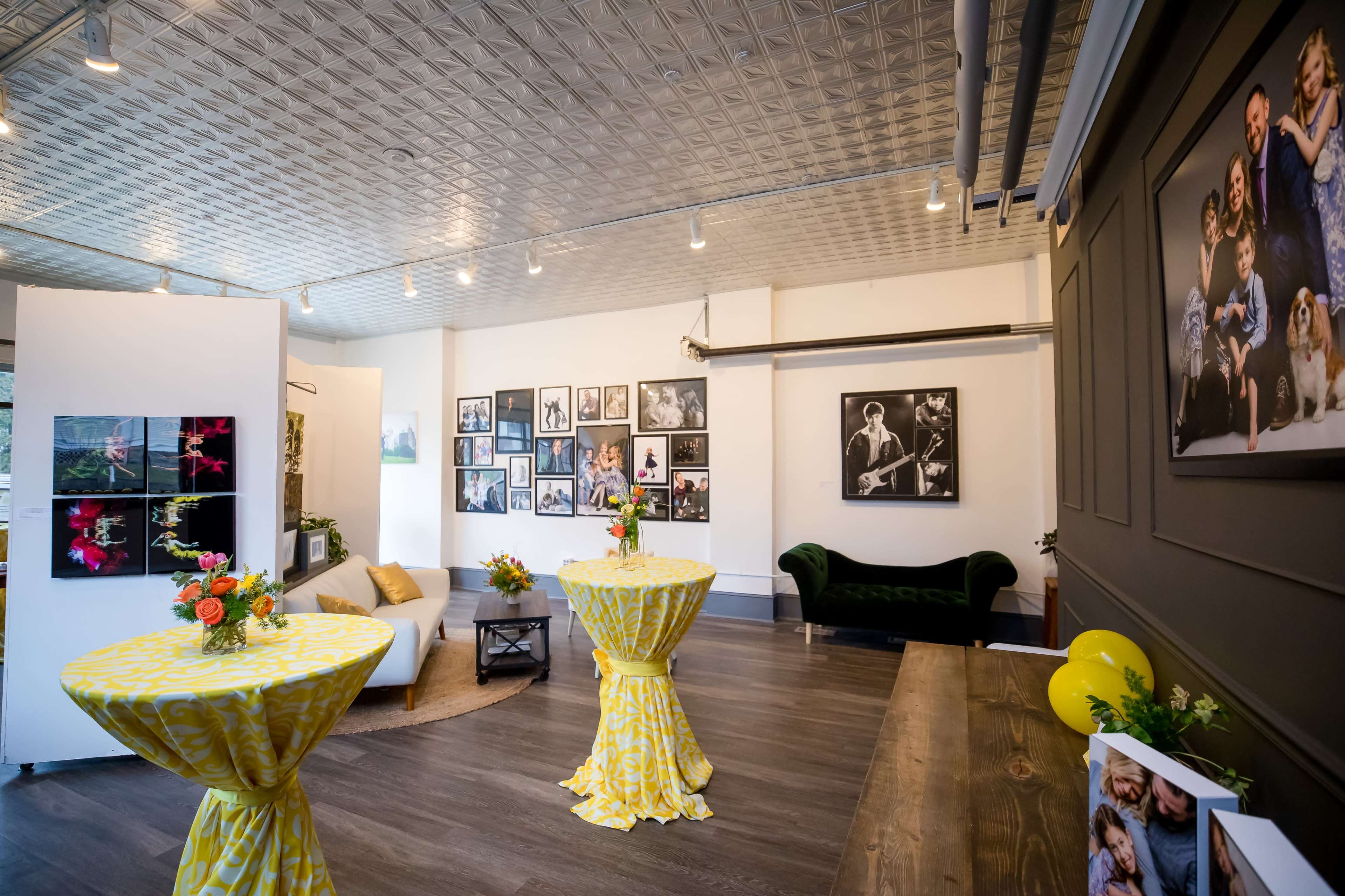 A spacious gallery with two yellow tables set with flowers, surrounded by framed photographs on the walls.