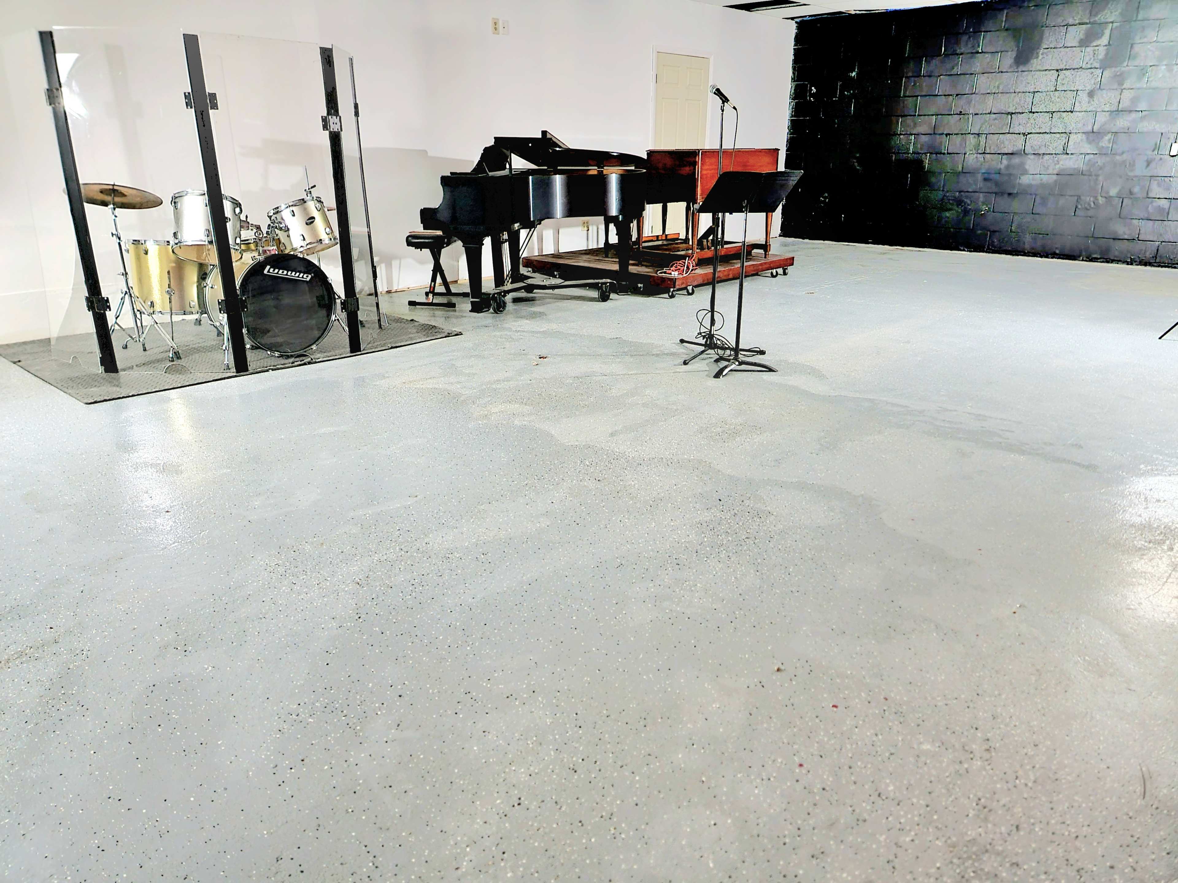 A music studio featuring a drum set, a piano, and a microphone stand on a polished concrete floor.